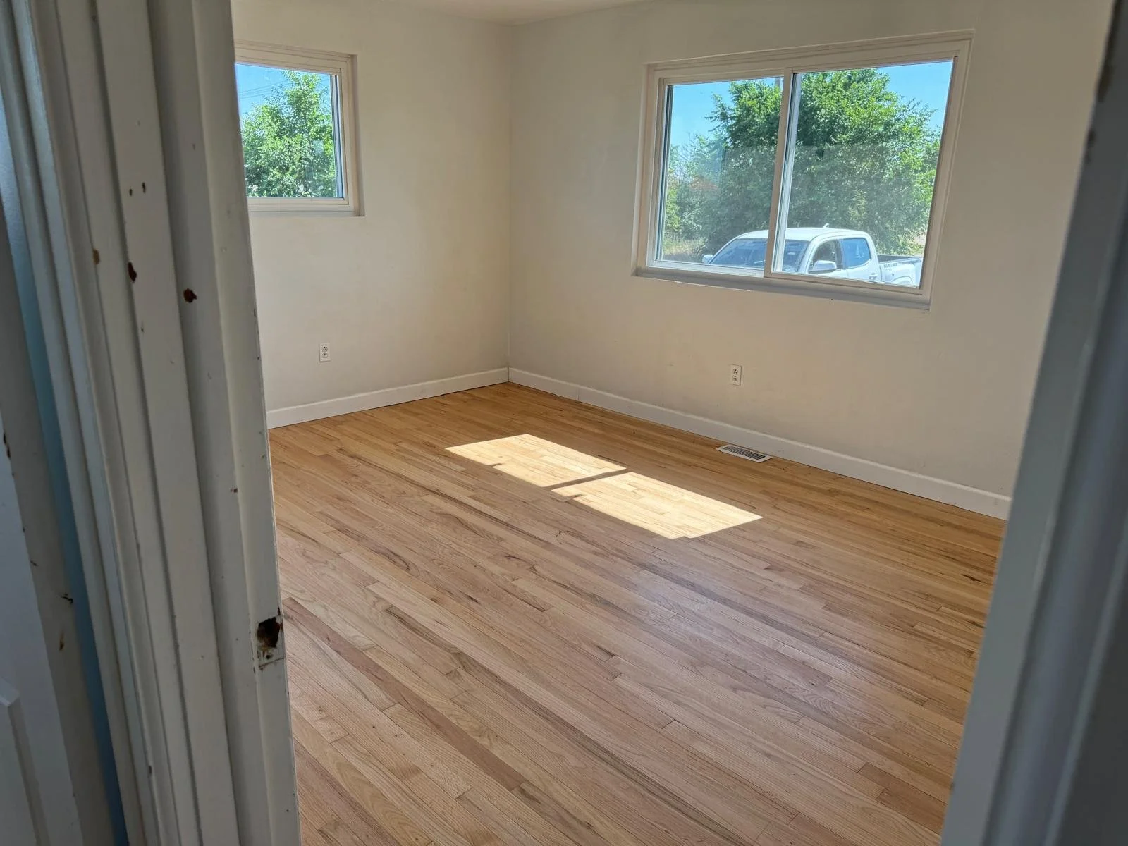 Empty room with hardwood floors, two windows letting in natural light, and white walls.
