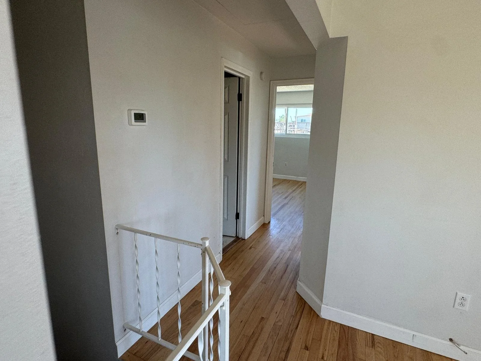 Interior view of a house hallway with wood flooring, white walls, open doorways, a thermostat on the wall, and a staircase with a white railing.