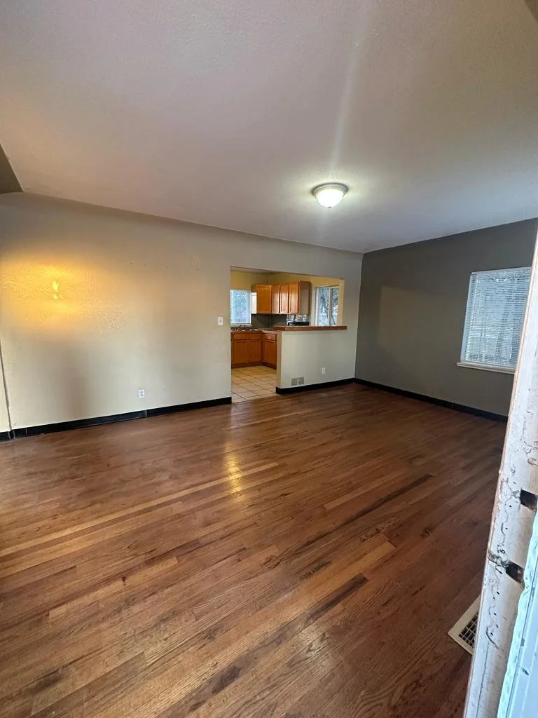 Empty living room with hardwood floors, beige and gray walls, ceiling light, and a view into the kitchen and window with blinds.