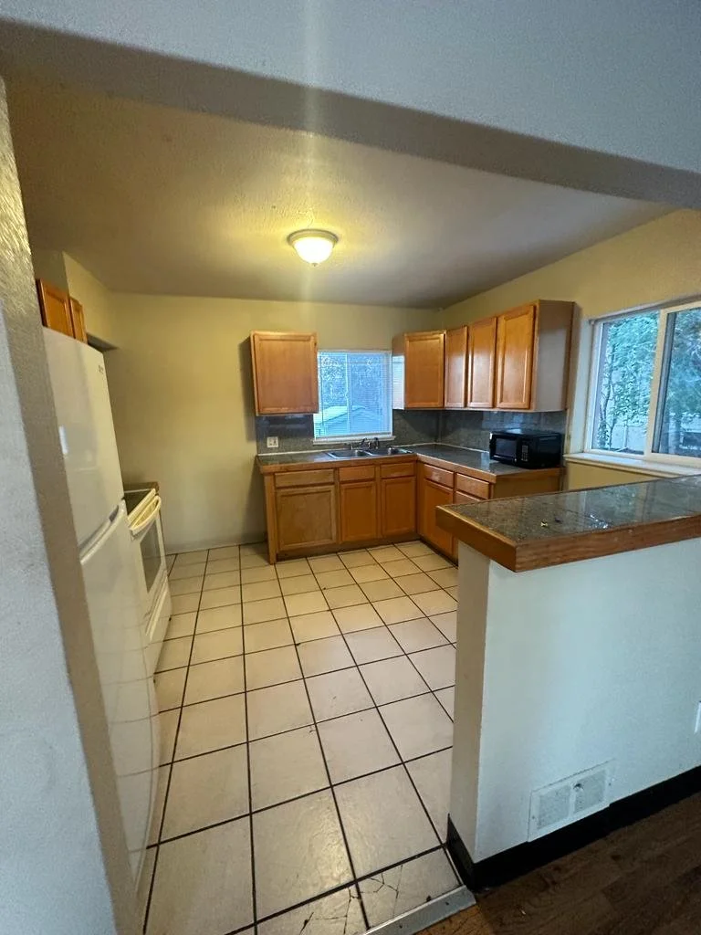 Empty kitchen with wooden cabinets, white appliances, dark countertop, tiled floor, and two windows letting in natural light.