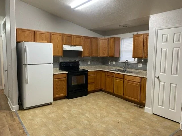 Empty kitchen with wooden cabinets, white refrigerator, black stove, and beige tile floor.