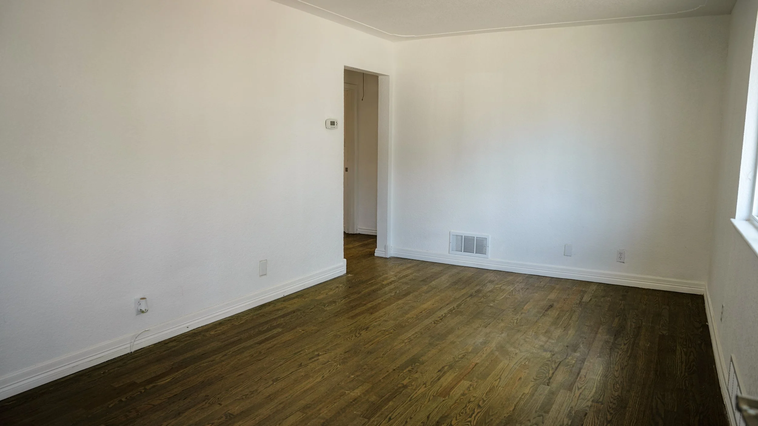 Empty room with white walls and hardwood floor, with a doorway leading to another room and a window on the right side.
