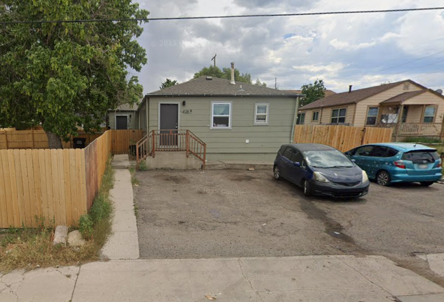 A small, single-story house with green siding and a gray roof. There's a wooden ramp and steps leading to the front door, with a wooden fence on either side. Two cars are parked in front of the house on a dirt parking area. The sky is cloudy, and neighboring houses are visible in the background.