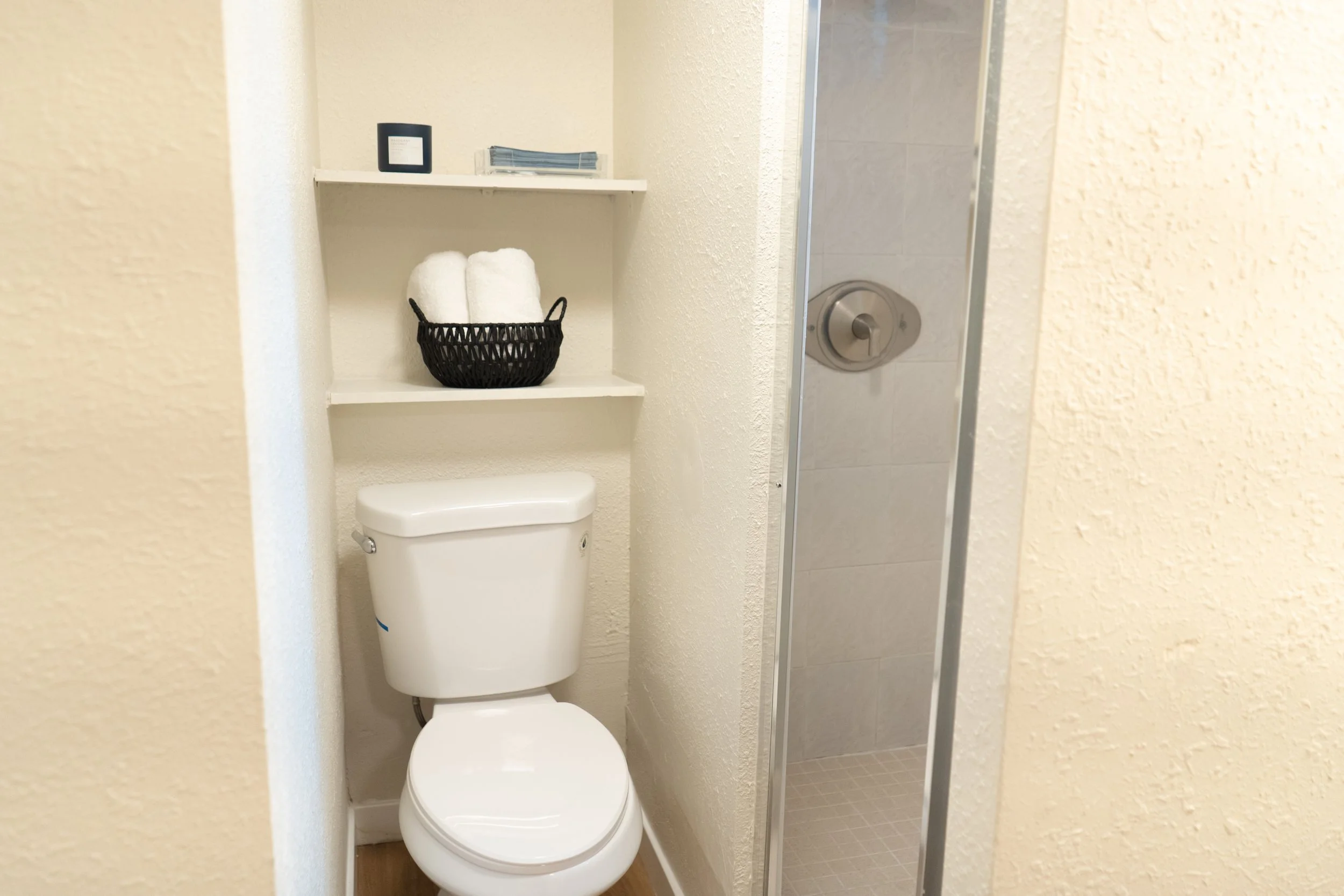 A small bathroom with a toilet, black basket with white towels on a shelf, and a shower with a glass door and metal handle.