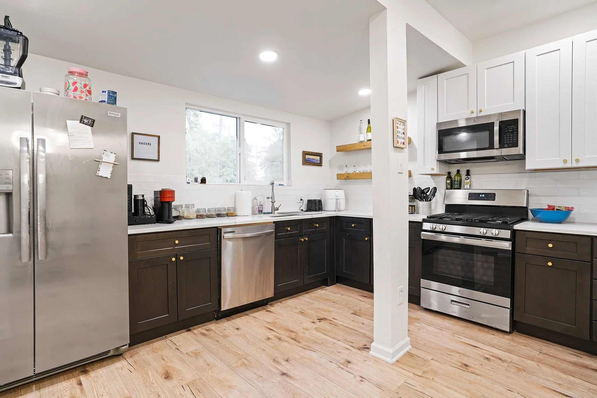 Modern kitchen with black lower cabinets, white upper cabinets, stainless steel appliances, wooden shelves, and a wood floor. There is a window above the sink, and various kitchen items are on the counters.