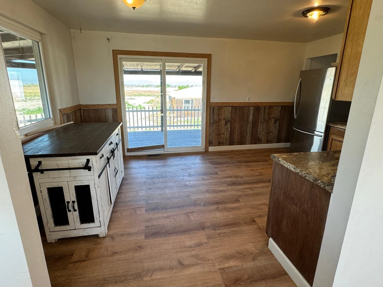 A kitchen with wooden flooring, a sliding glass door leading to an outdoor deck, a white cabinet with black hardware, granite countertops, and a stainless steel refrigerator. Wood paneling decorates the walls.