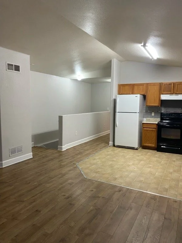 Empty kitchen and living area with white walls, wood flooring, a white refrigerator, and black stove. Kitchen has wooden cabinets and a beige tiled floor section.