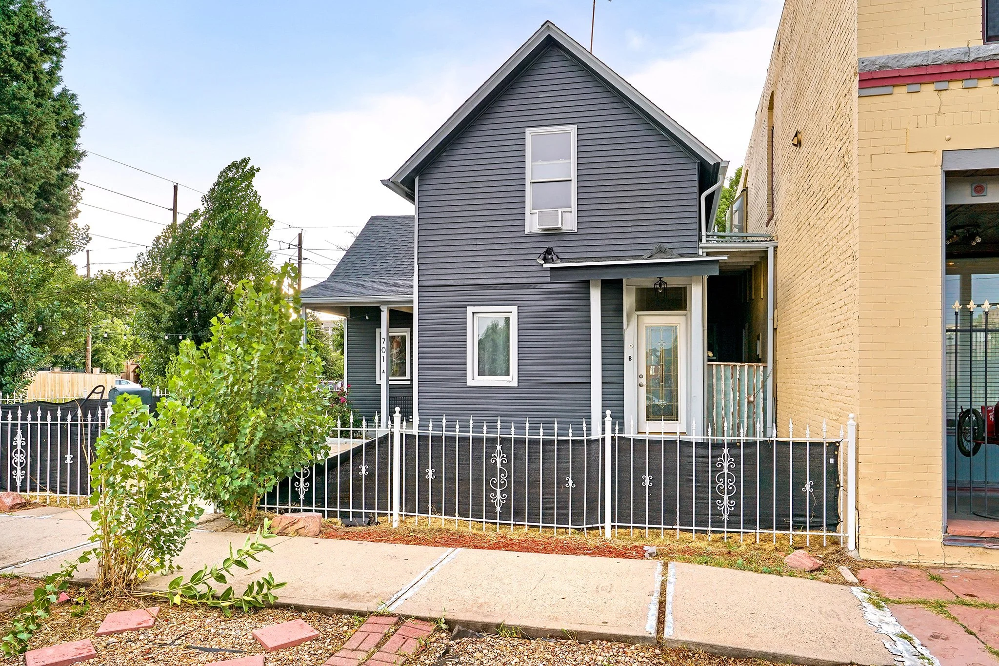 A blue two-story house with white trim and a small front porch, located between two other buildings. The house has a black fence and a small landscaped area with a bush in front.