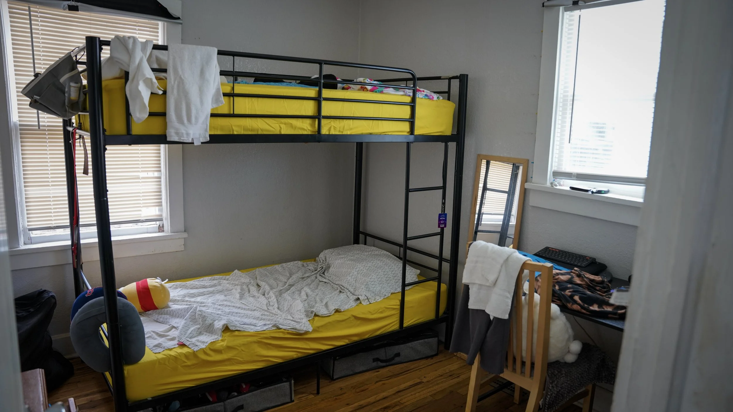 A bedroom with a metal bunk bed that has yellow mattresses, the lower bed is unmade with rumpled white sheets, a neck pillow, and a plush toy. There is a window with blinds on the left, a small mirror leaning against the wall, and another window on t