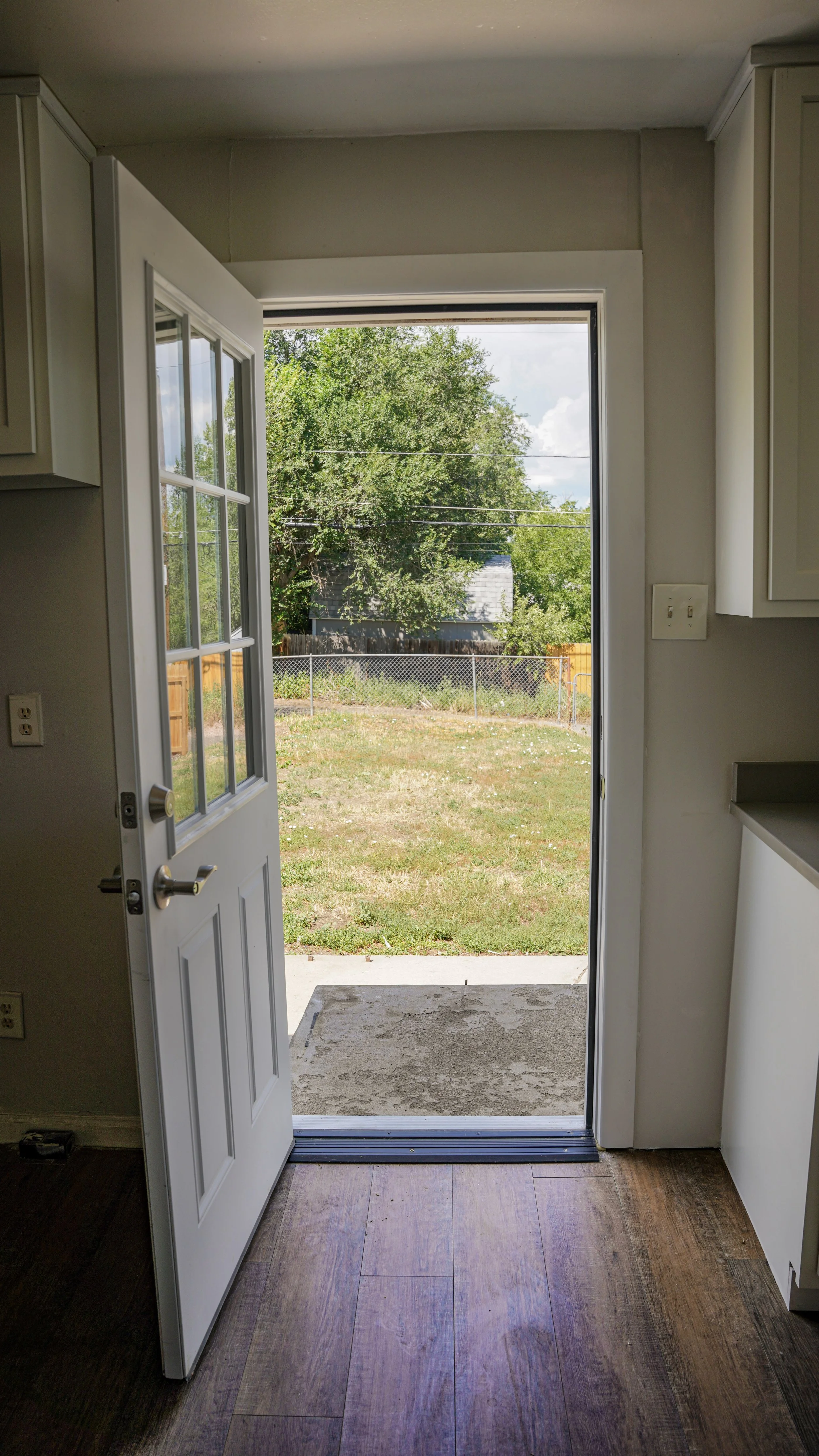 Open back door leading to a backyard with grass, a fence, and trees, taken from inside a kitchen with hardwood floors and white cabinets.