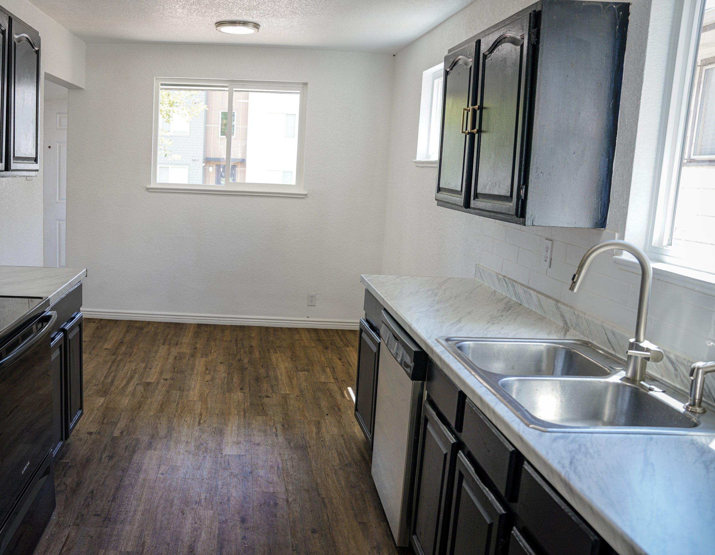 Empty kitchen with white walls, wooden floor, black cabinets, marble countertop, stainless steel sink, and two windows.