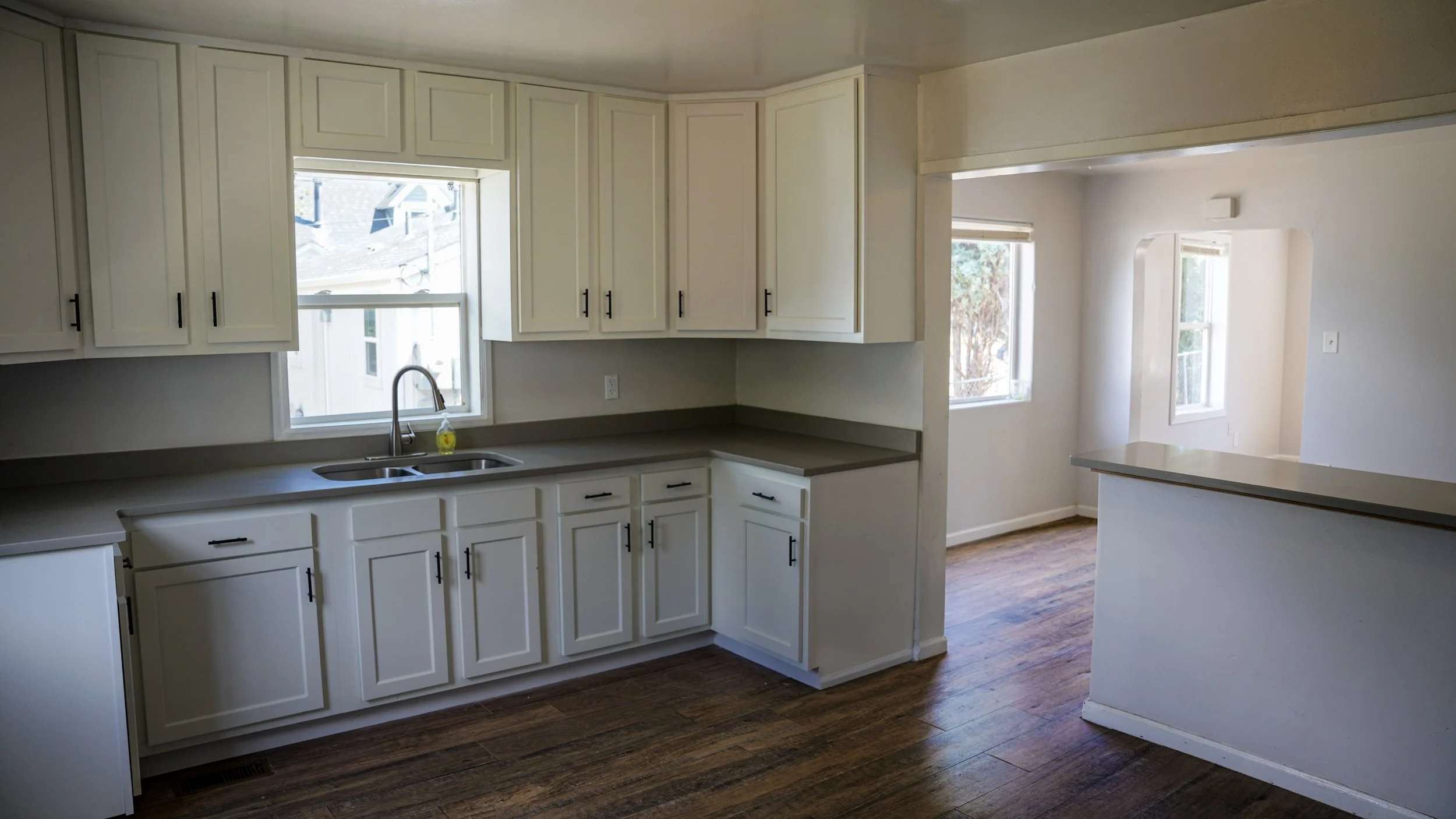 Kitchen with white cabinets, gray countertops, a double sink, on a wood floor, with windows letting in natural light.