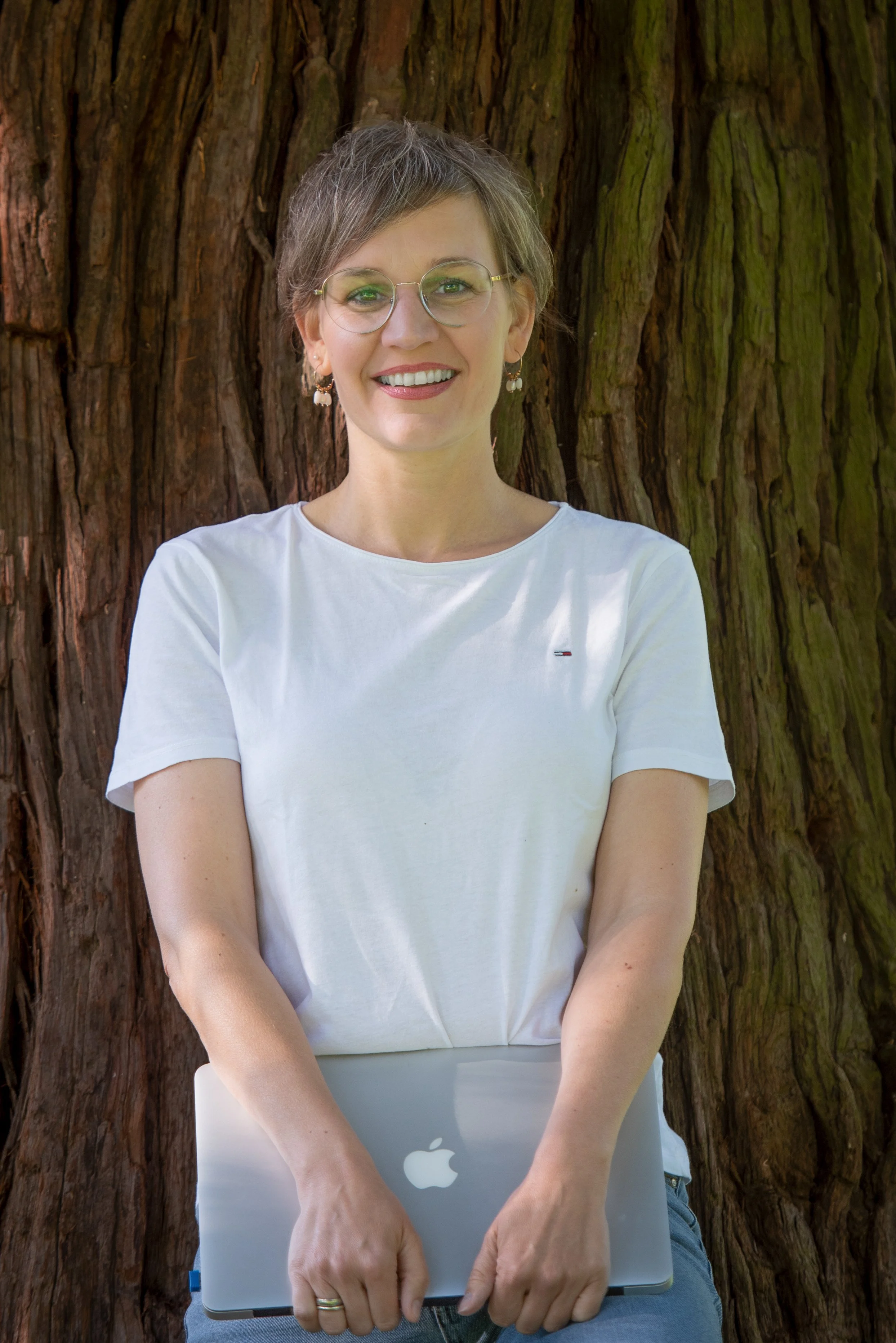 Frau mit Brille hält einen MacBook vor sich, steht vor einem Baumstamm im Freien, trägt ein weißes T-Shirt und lächelt.