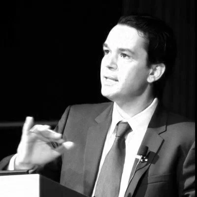 Black and white photo of a man in a suit speaking at a podium with a microphone, gesturing with his hand.