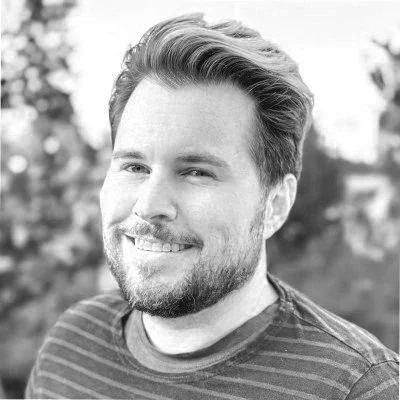 Black and white photo of a smiling man with styled hair and a beard, wearing a striped shirt, outdoors with trees in the background.