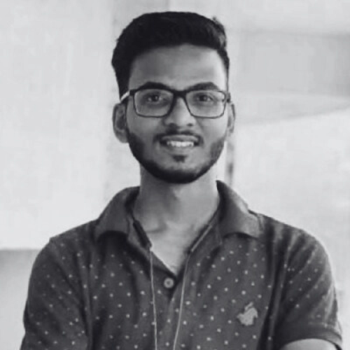 A young man with glasses and a beard smiling, wearing a collared shirt with small polka dots.