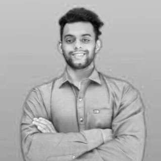 Black and white photo of a young man with dark hair and a beard, smiling, arms crossed, wearing a button-up shirt.