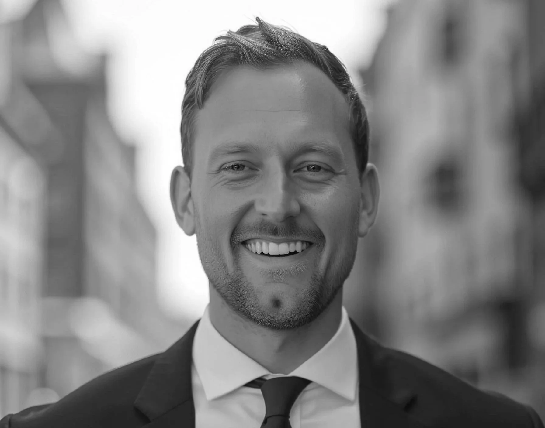 A smiling man in a suit, outdoors in an urban setting, black and white photograph.