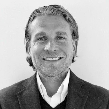 A black-and-white headshot of a smiling man with light-colored hair, wearing a suit jacket and a collared shirt.