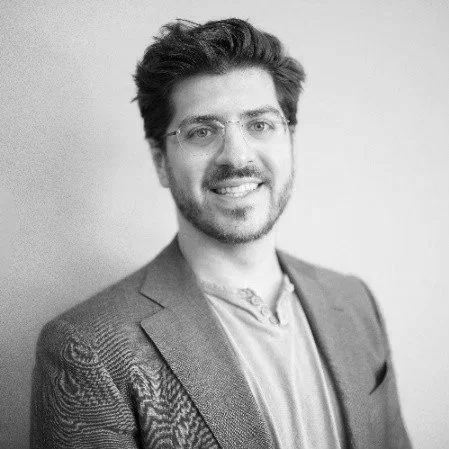 Black and white portrait of a young man with glasses, beard, smiling, wearing a blazer and shirt, standing against a plain background.