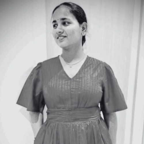 A woman smiling, wearing a dress with puffed sleeves, standing indoors against a plain wall.
