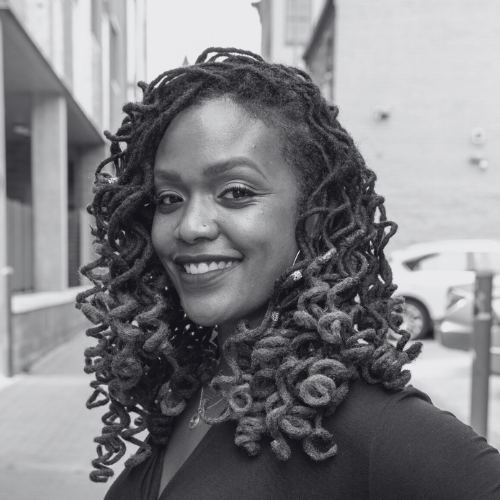 A woman with dark curly dreadlocks smiling outdoors.