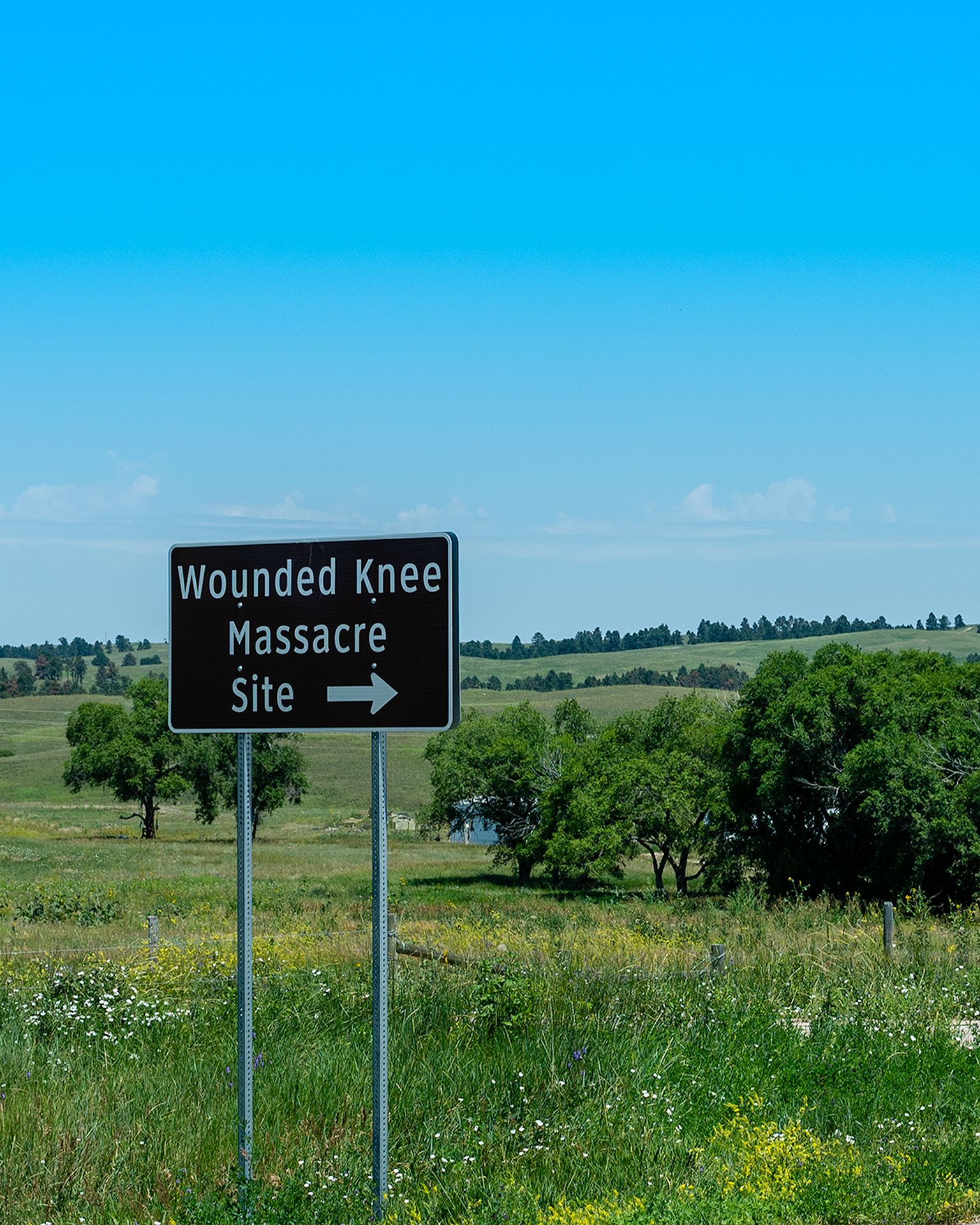 Road sign indicating the site of a Wounded Knee massacre with a landscape of green hills, trees, and a blue sky in the background.