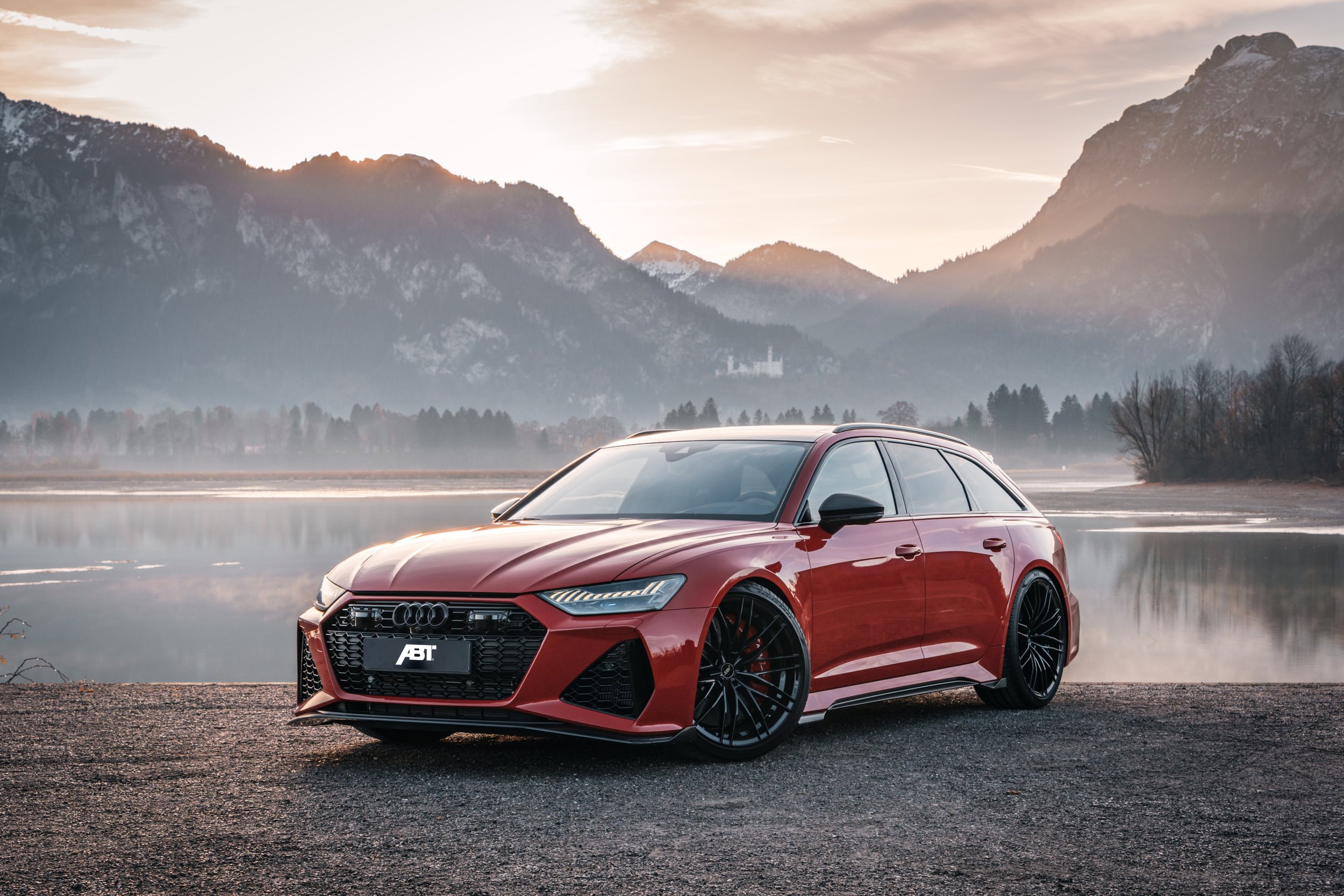 Red Audi station wagon parked near a lake with mountains in the background during sunset.