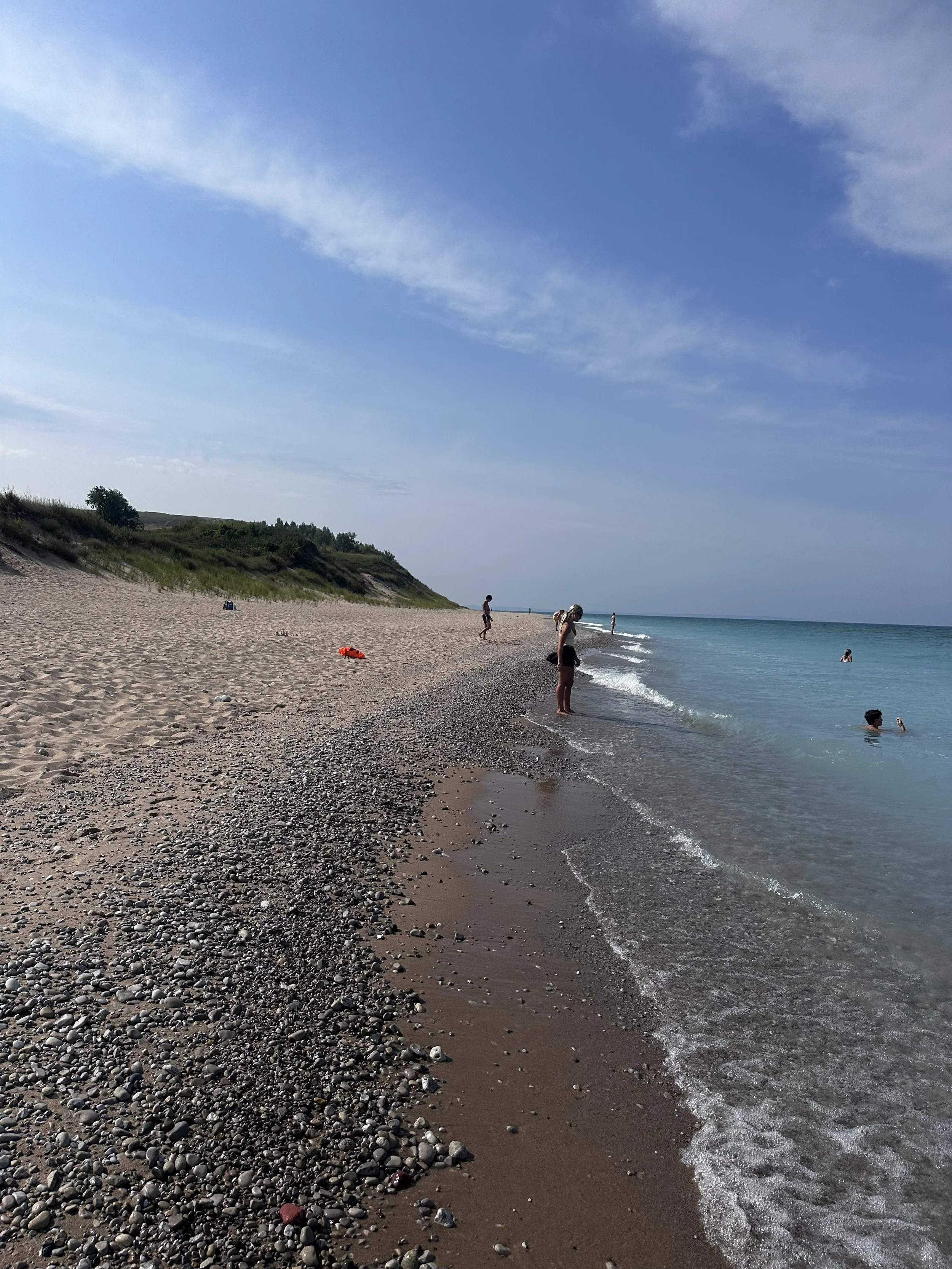 People enjoy a day at the beach with a sandy and pebbly shoreline, gentle waves, a partly cloudy sky, and some greenery on a hill in the background.