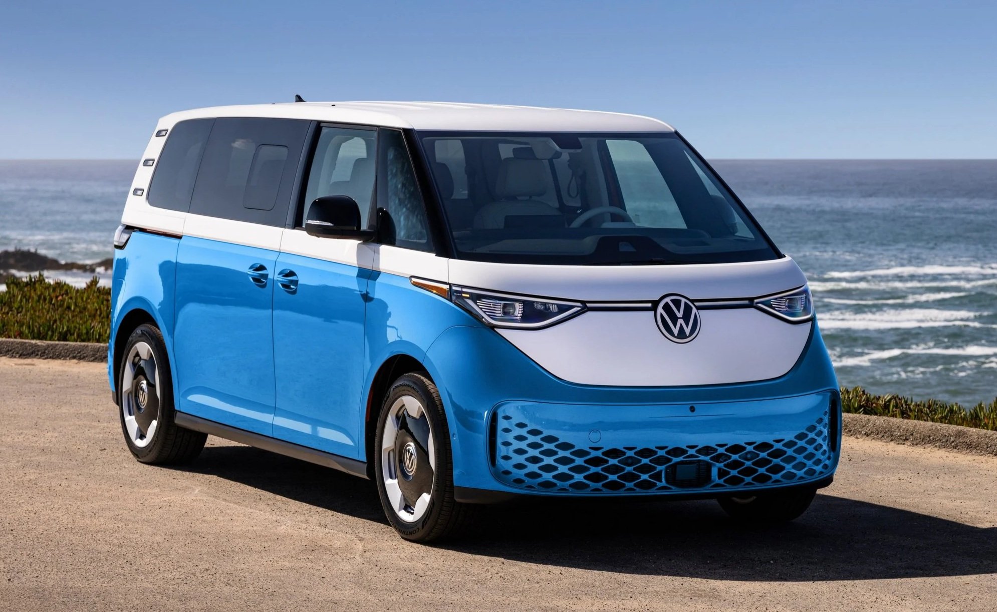 Electric Volkswagen ID. Buzz van parked on coastal road with ocean and blue sky in background.