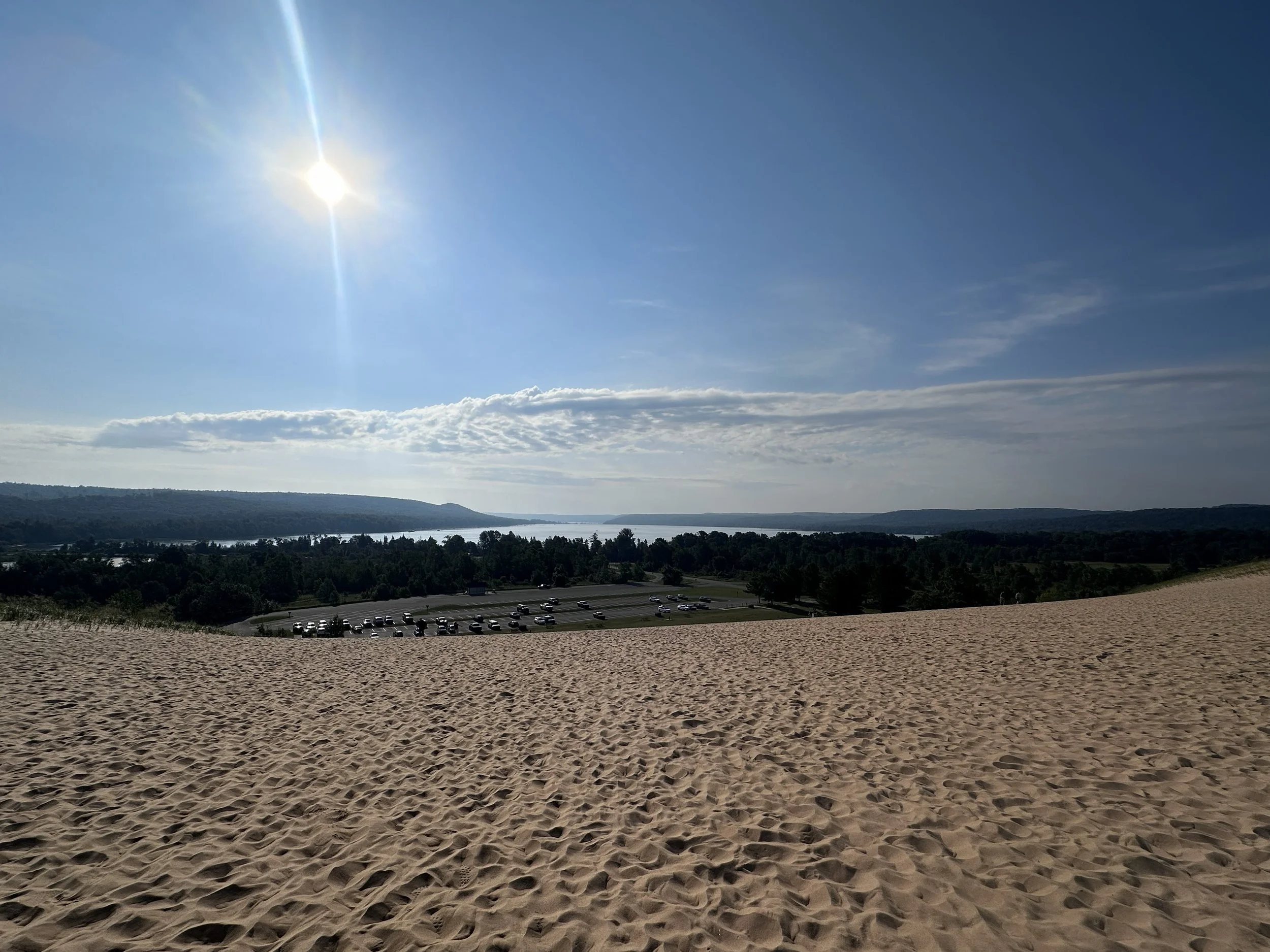 Sand dune leading to a parking lot with trees, water, and hills in distance under a clear blue sky with sunlight.