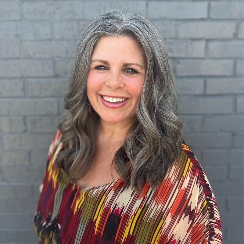 A smiling woman with long, wavy gray hair standing in front of a gray brick wall, wearing a colorful patterned blouse.