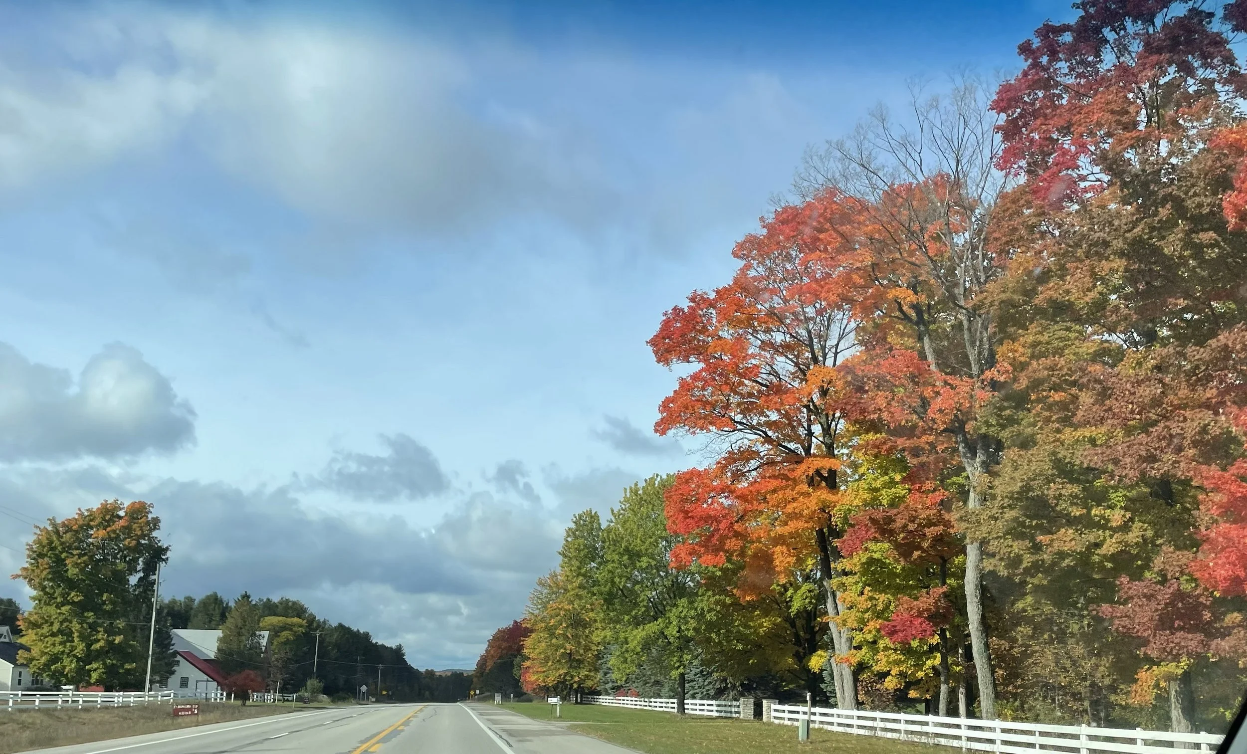 A scenic rural road lined with colorful autumn trees on the right side, with a white fence and a house in the distance, under a partly cloudy sky.