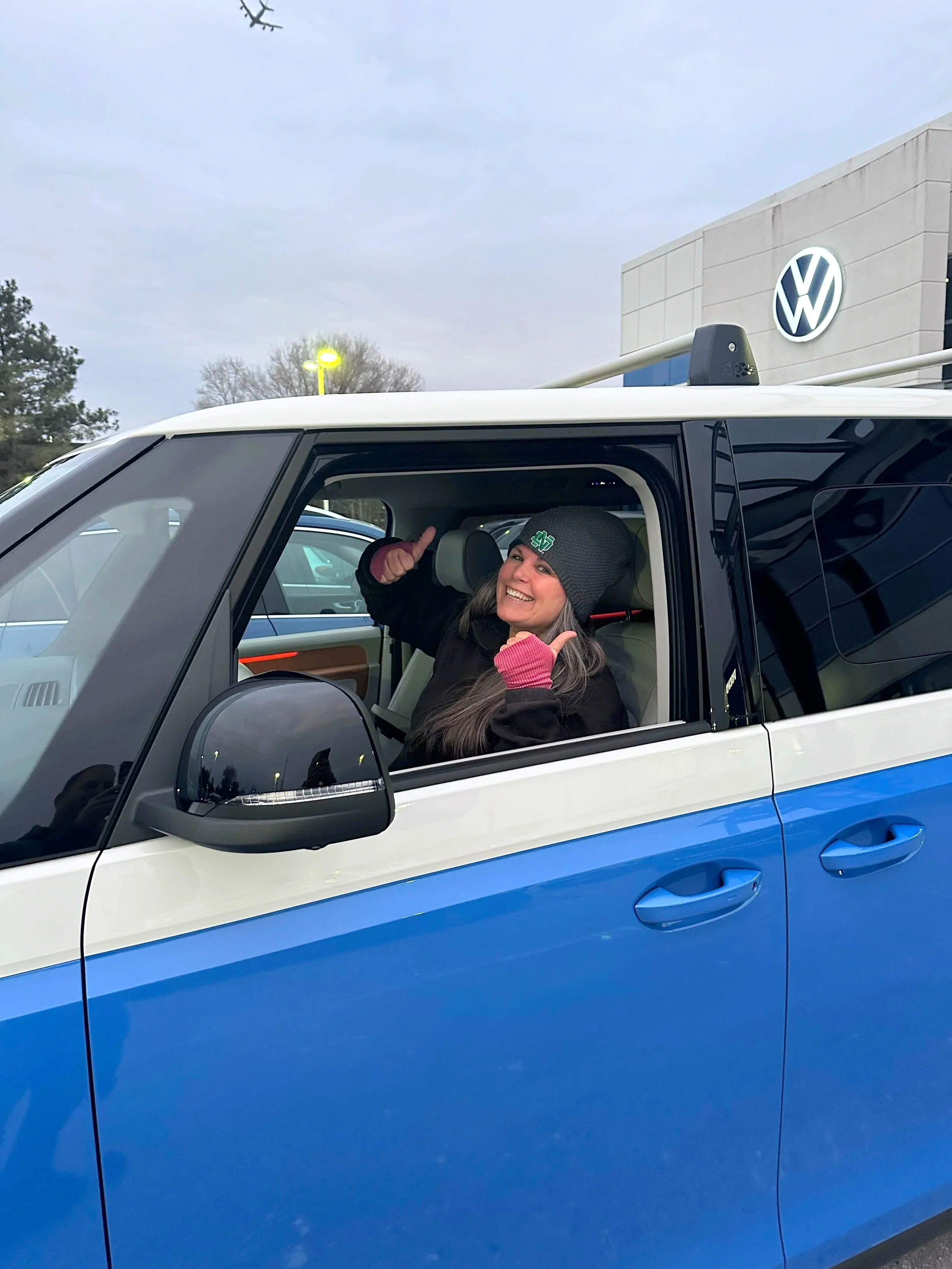 A woman sitting in the driver's seat of a blue and white vehicle, smiling and making a thumbs-up gesture with both hands, outside a Volkswagen dealership.