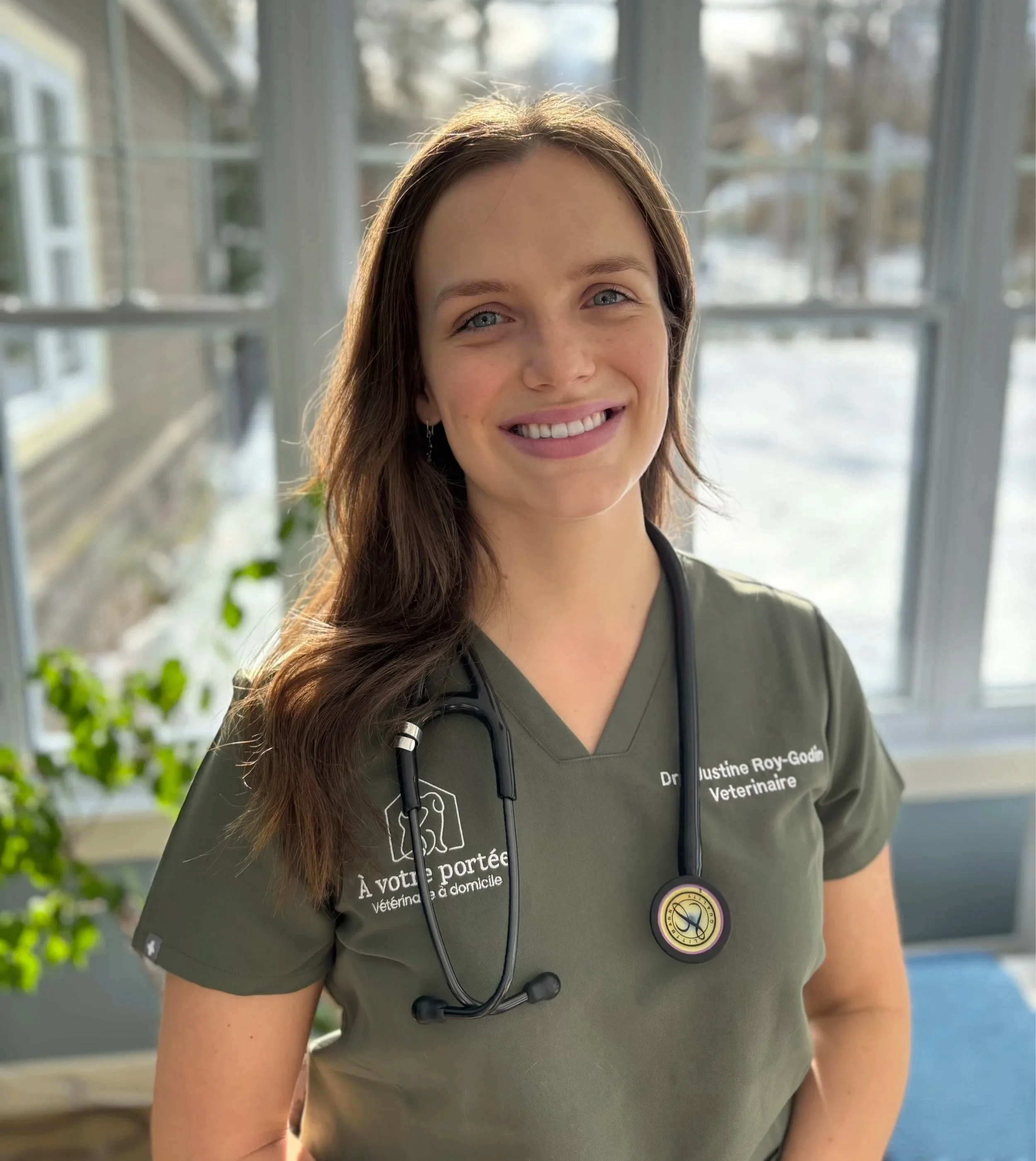 Une vétérinaire souriante porte une chemise verte avec une stéthoscope autour du cou, posée devant une fenêtre avec vue sur un jardin.