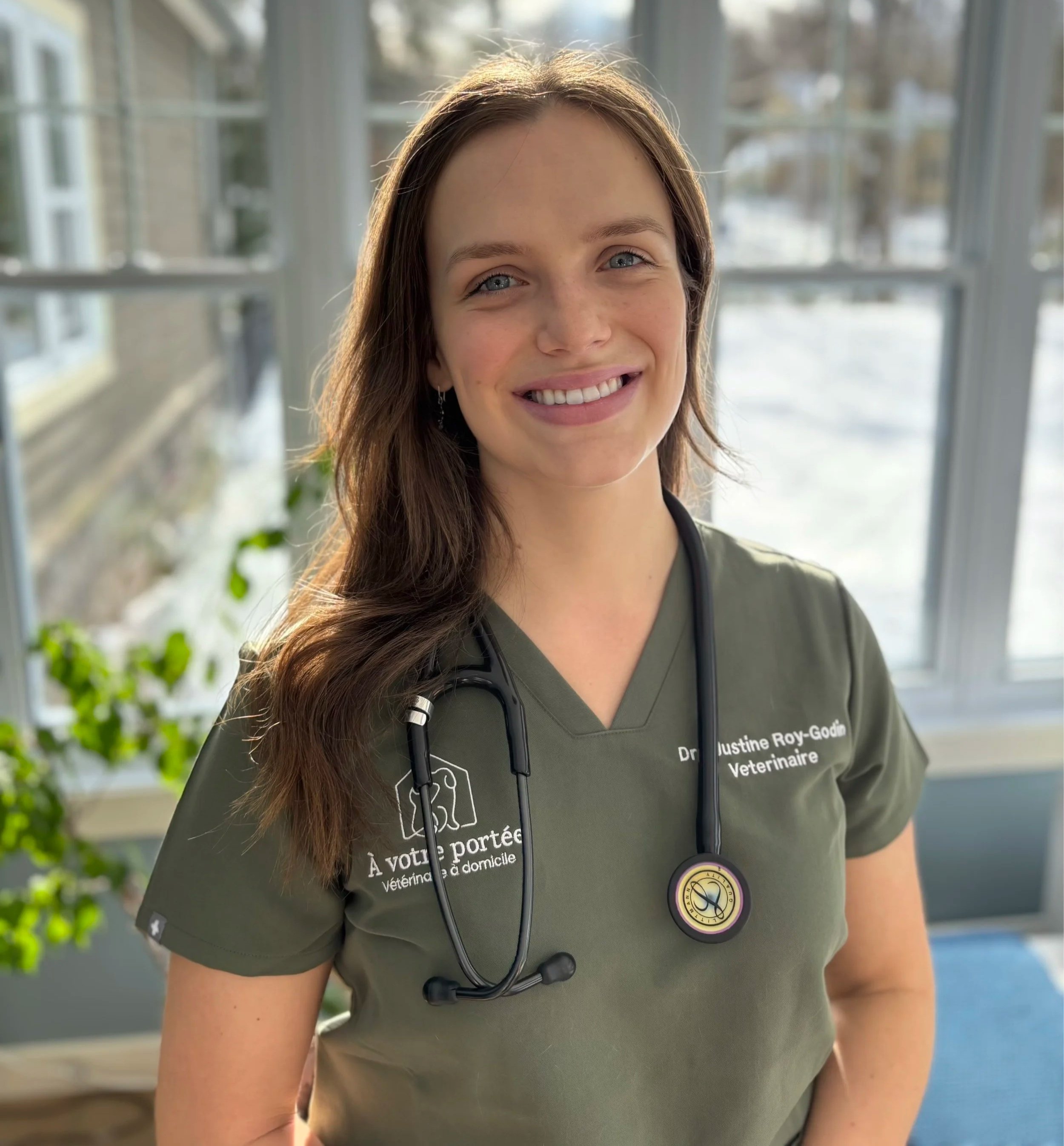 Une vétérinaire souriante portant un uniforme vert olive avec un stéthoscope autour du cou, devant une fenêtre avec vue sur l'extérieur en hiver.