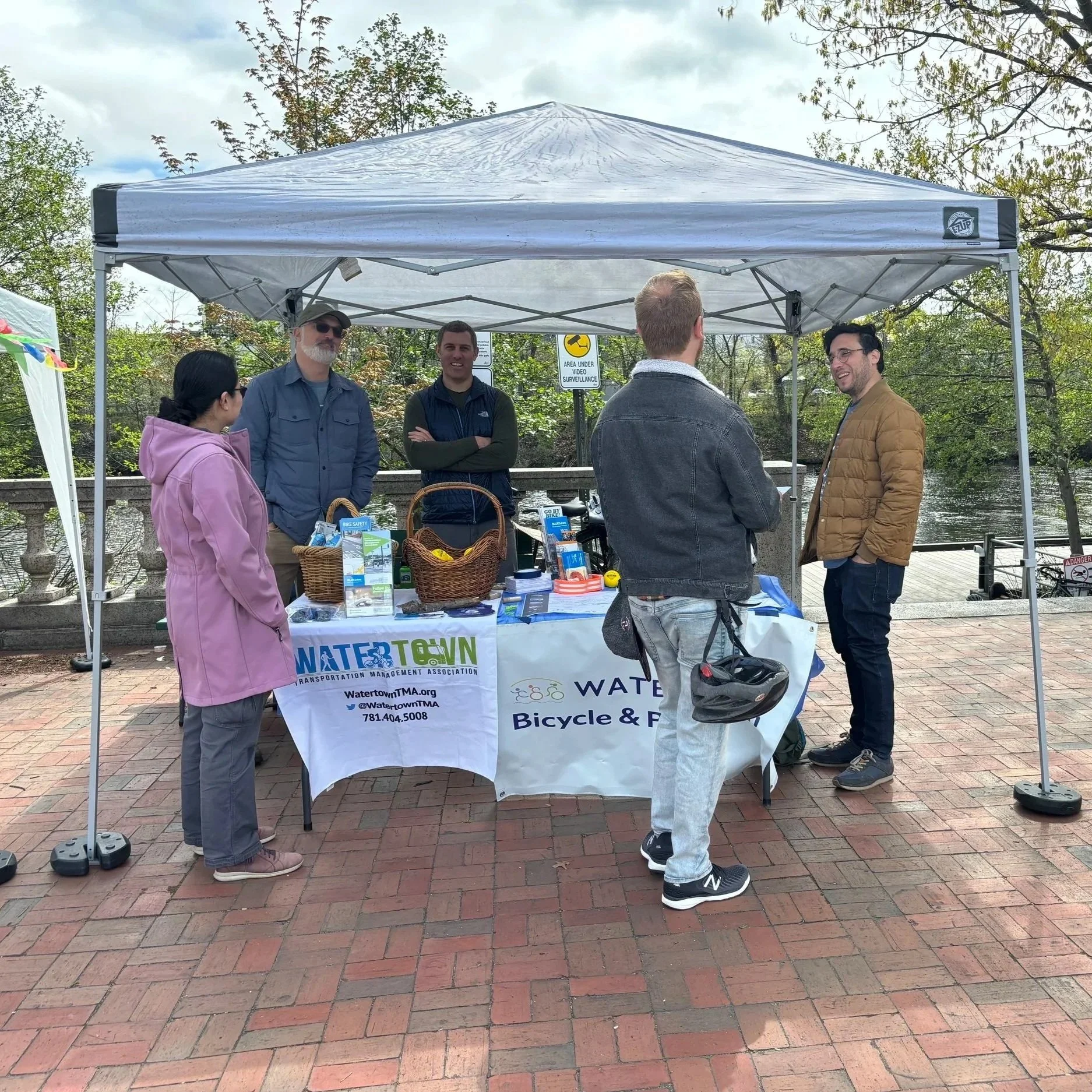 Group of five people standing under a white canopy tent, engaged in conversation, with informational pamphlets and baskets on a table, outdoors near a river or pond with trees in the background.