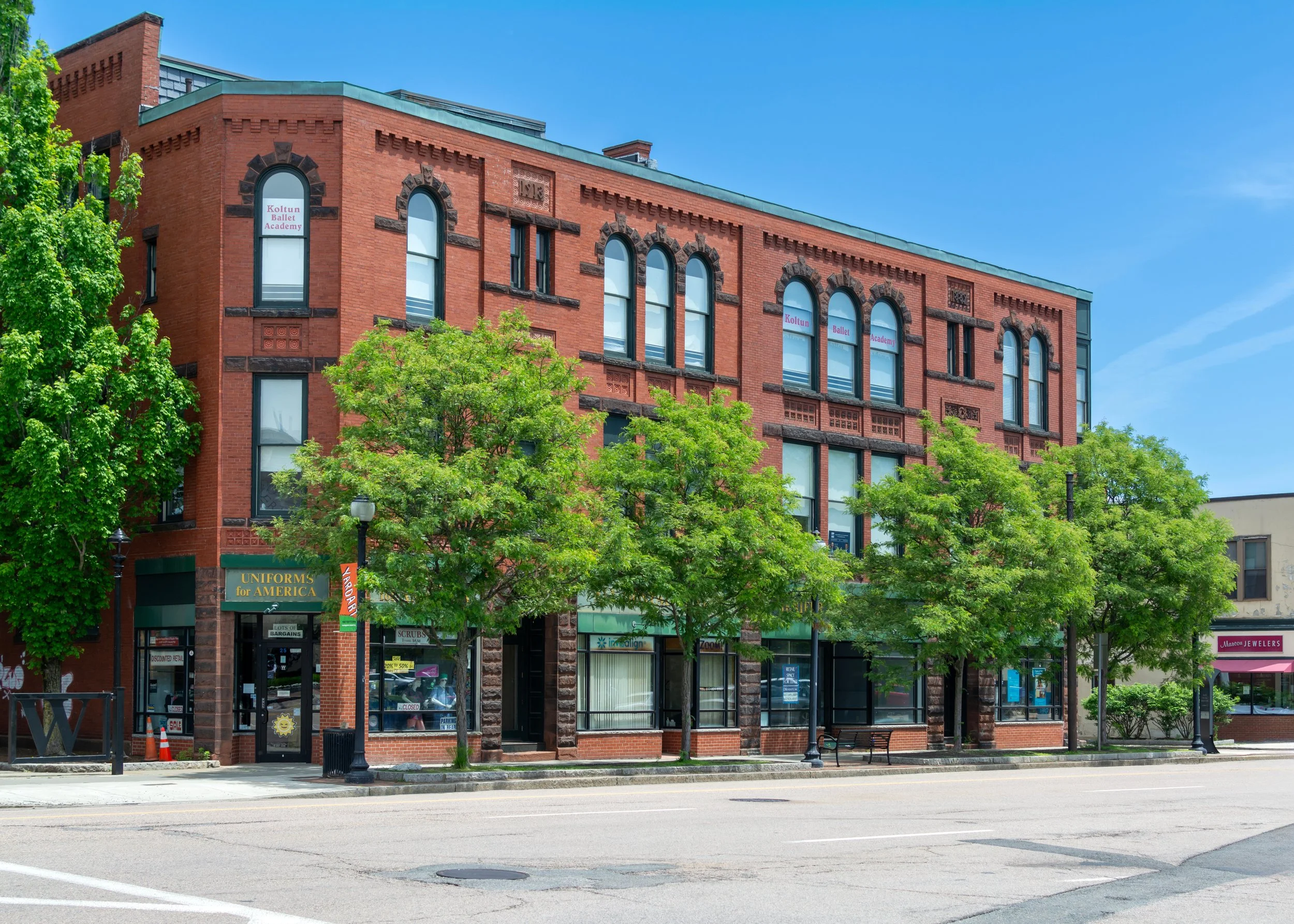 A three-story red brick building with trees in front, storefronts on the ground level, and signs indicating a ballet academy and a jewelers. Clear blue sky in the background.