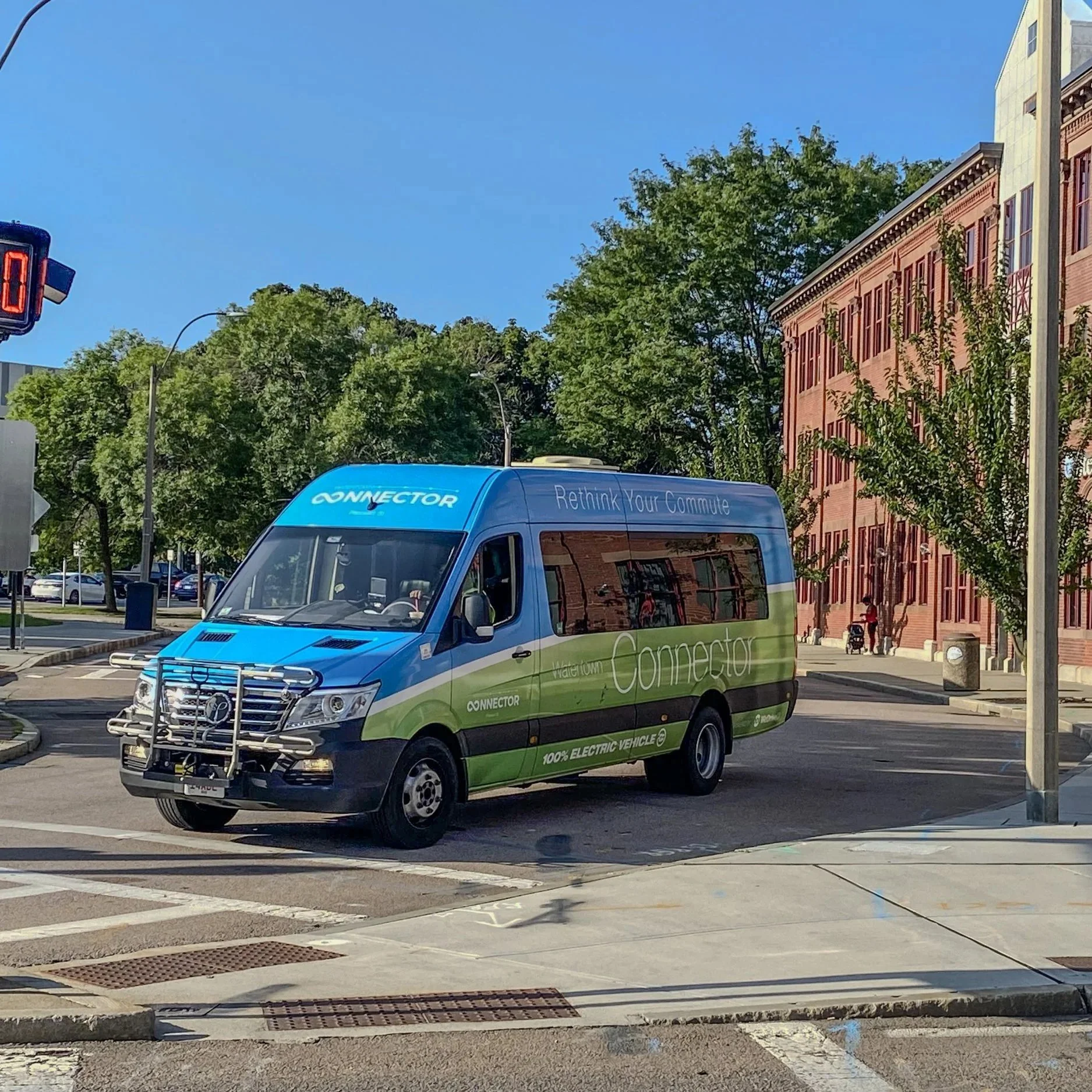 Electric shuttle bus with blue and green color scheme, labeled 'Connector' and 'Rethink Your Commute,' parked on city street in front of brick building with 'Stewart' sign.
