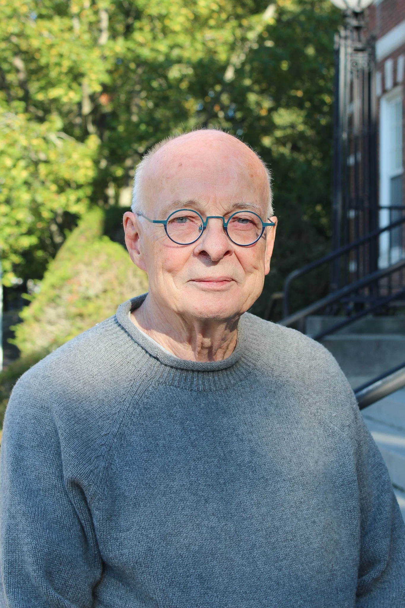 An elderly man with glasses, wearing a gray sweater, standing outdoors in front of green trees and a building with stairs.