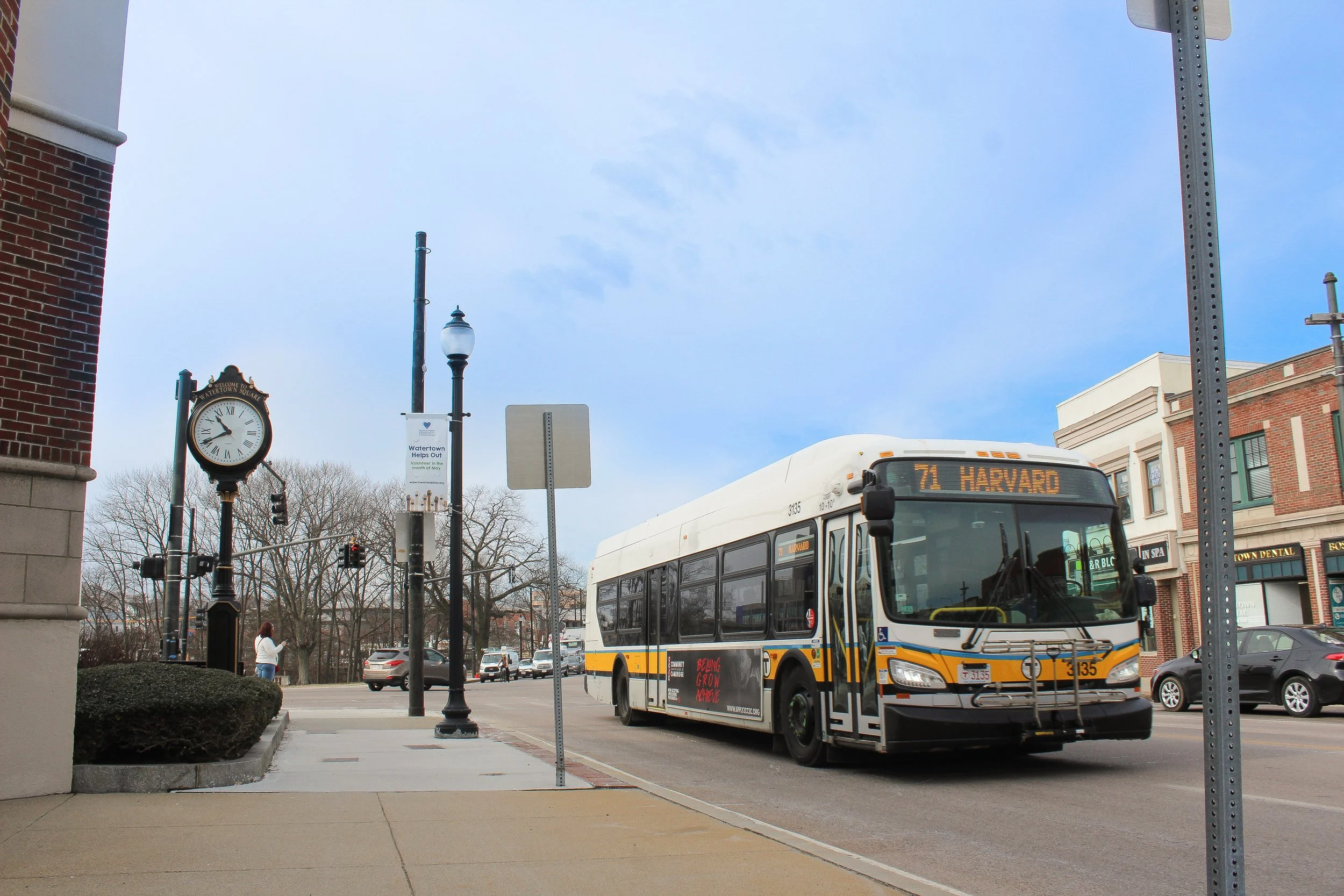 A city street scene with a white and yellow bus labeled '71 HARVARD,' parked on the side of the road. There are young trees without leaves, a street clock, and a woman walking on the sidewalk. Buildings with storefronts and parked cars are visible in the background, and the sky is partly cloudy.