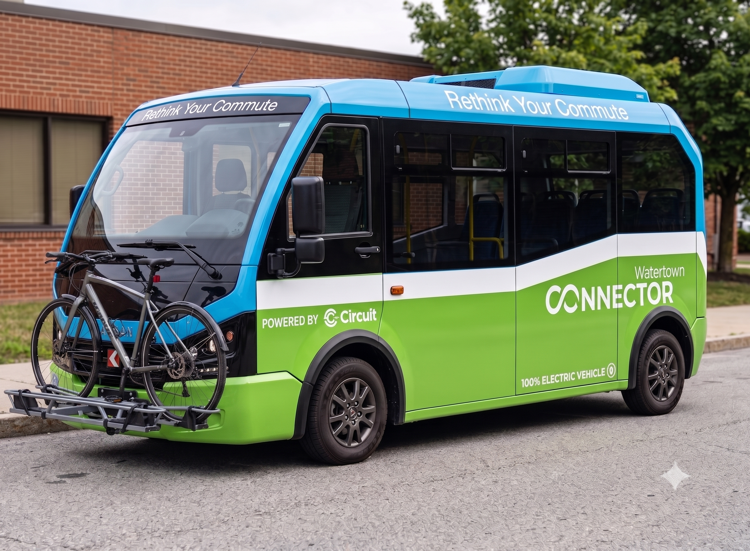 Electric shuttle bus with blue and green color scheme, labeled 'Connector' and 'Rethink Your Commute,' parked on city street in front of brick building with 'Stewart' sign.