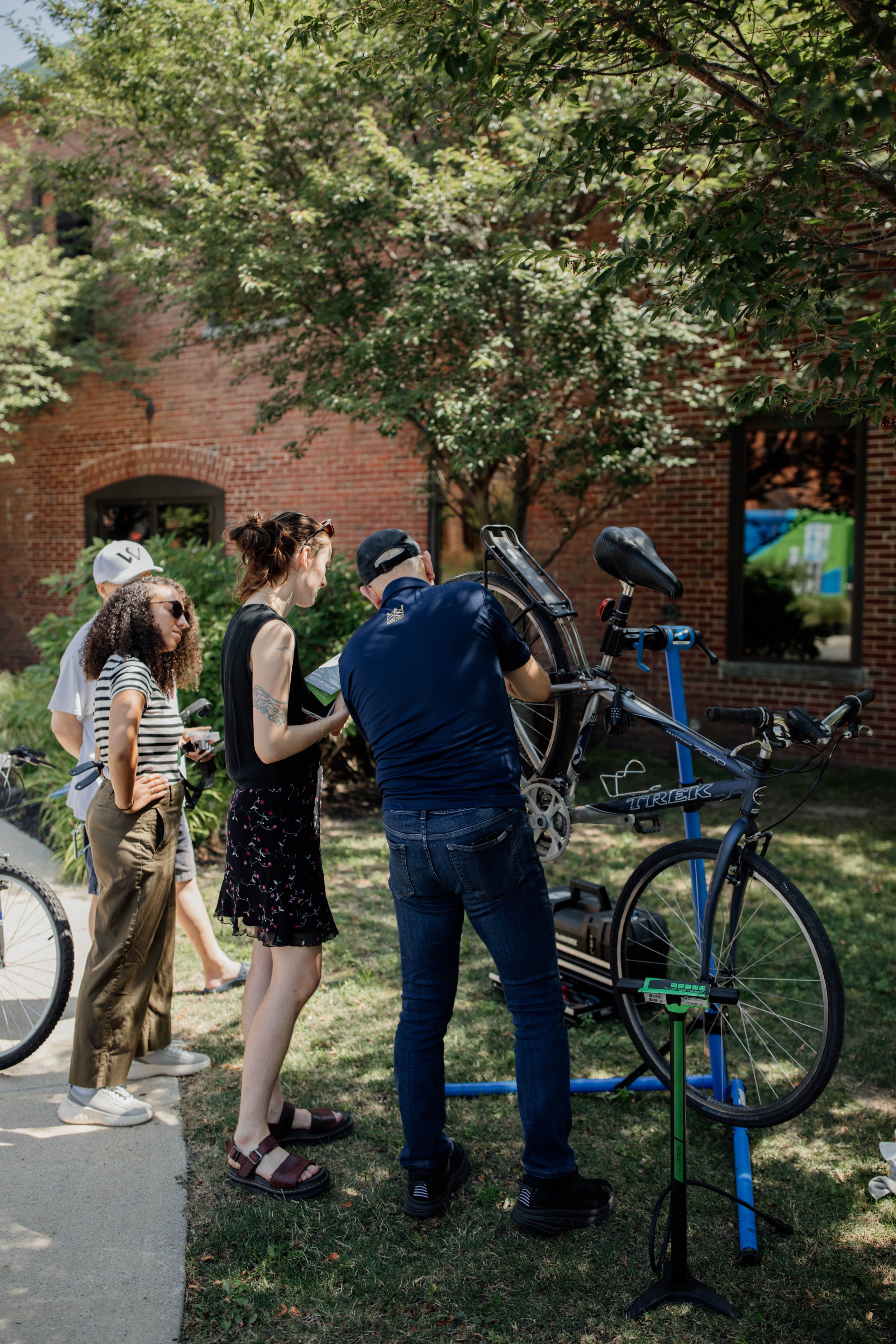 A group of five people, including three women and two men, gathered outside on the grass viewing a bicycle being worked on by a man. The man is using tools on the bicycle which is mounted on a repair stand, with a digital scale nearby.