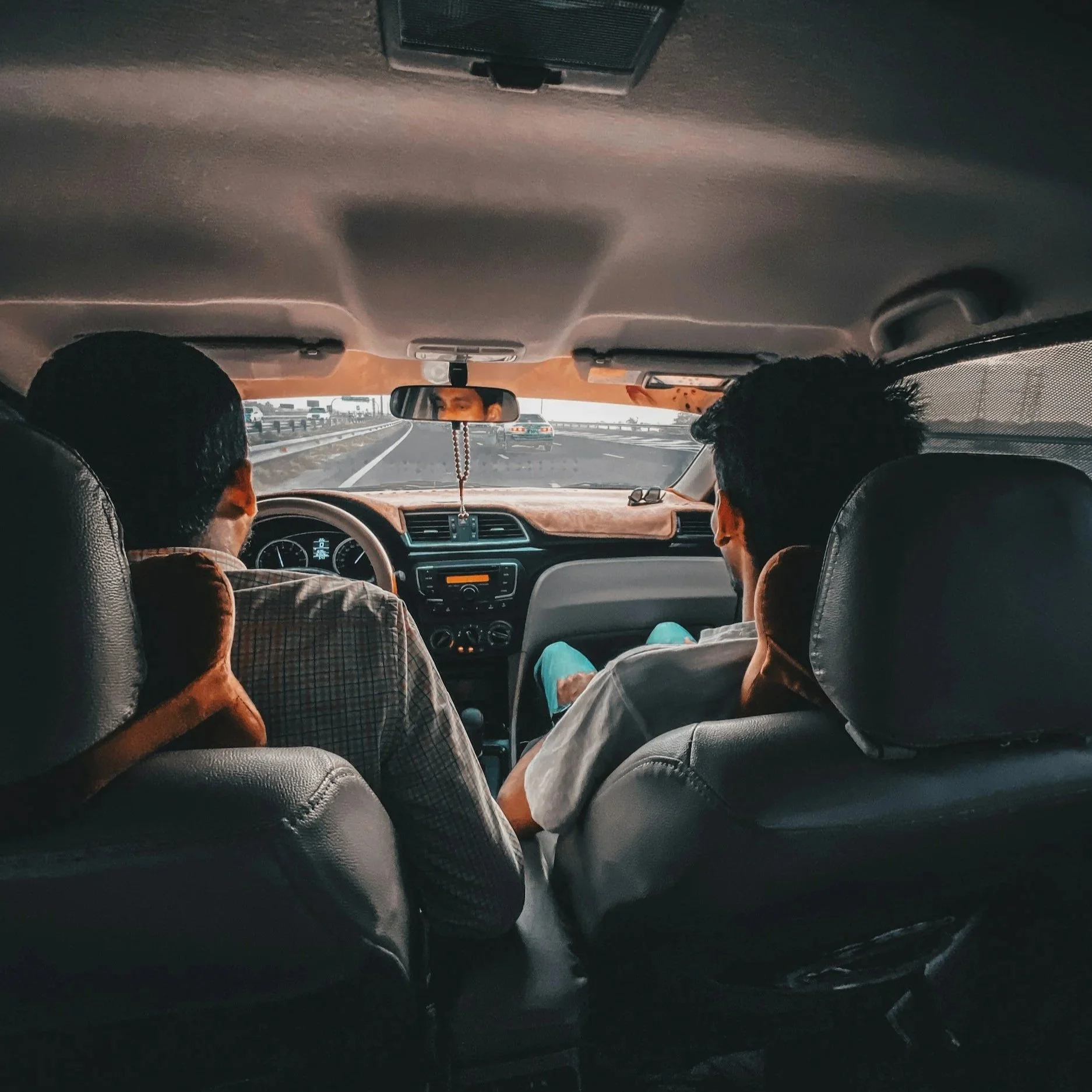 View from inside a car showing the dashboard, rearview mirror, and highway traffic, with two passengers visible from behind.