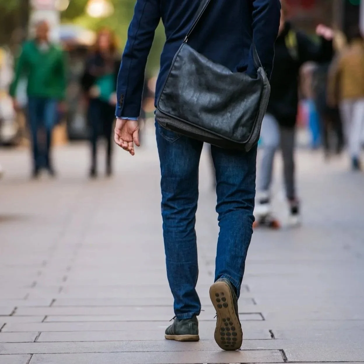 Person wearing a blue jacket and jeans, carrying a black shoulder bag, walking on a paved sidewalk with other pedestrians in the background.