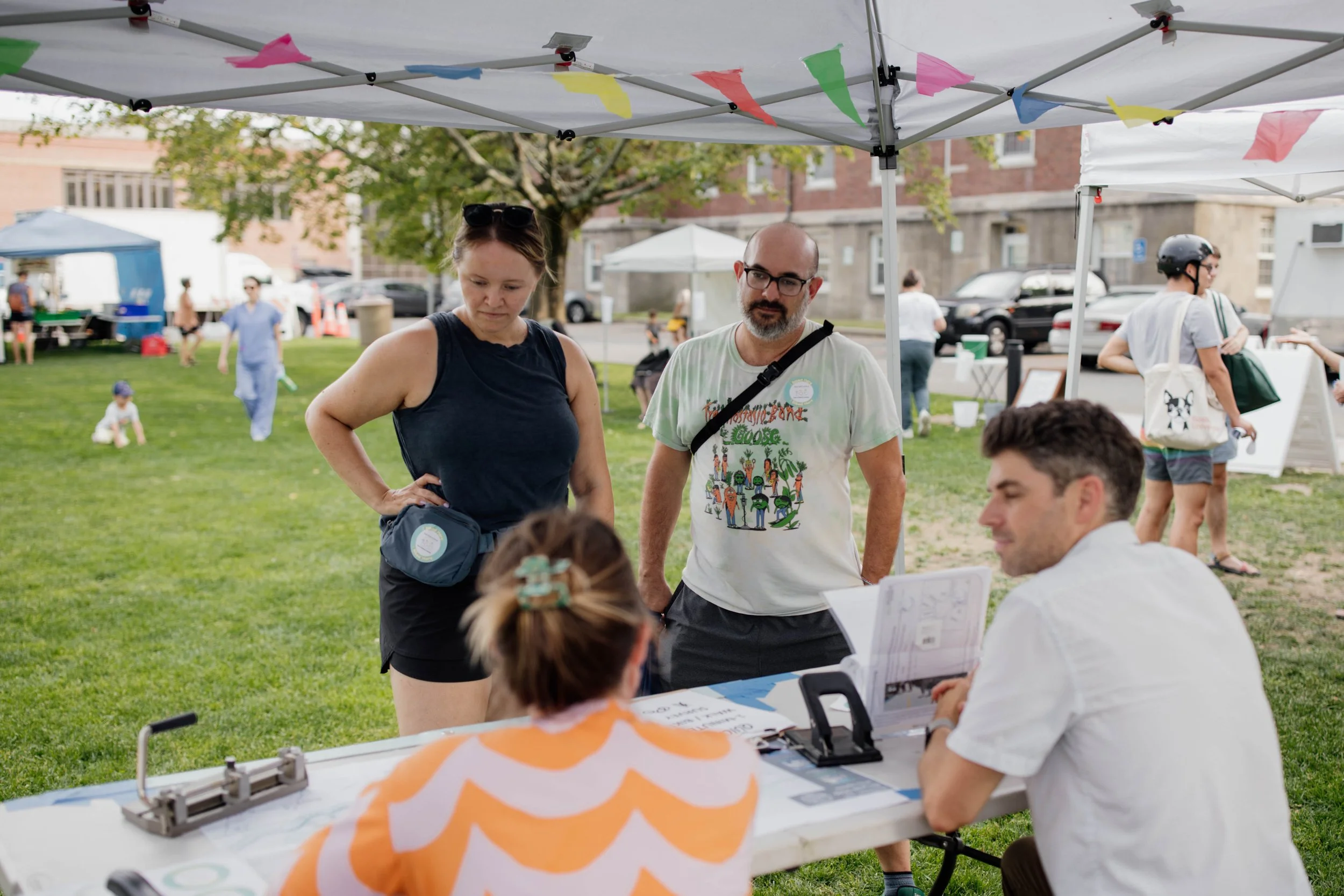 People at an outdoor event under a white canopy, with a grassy area and other tents in the background. Three individuals are standing and talking to a man seated at a table, which has papers and a sign. The scene appears to be a community fair or festival.