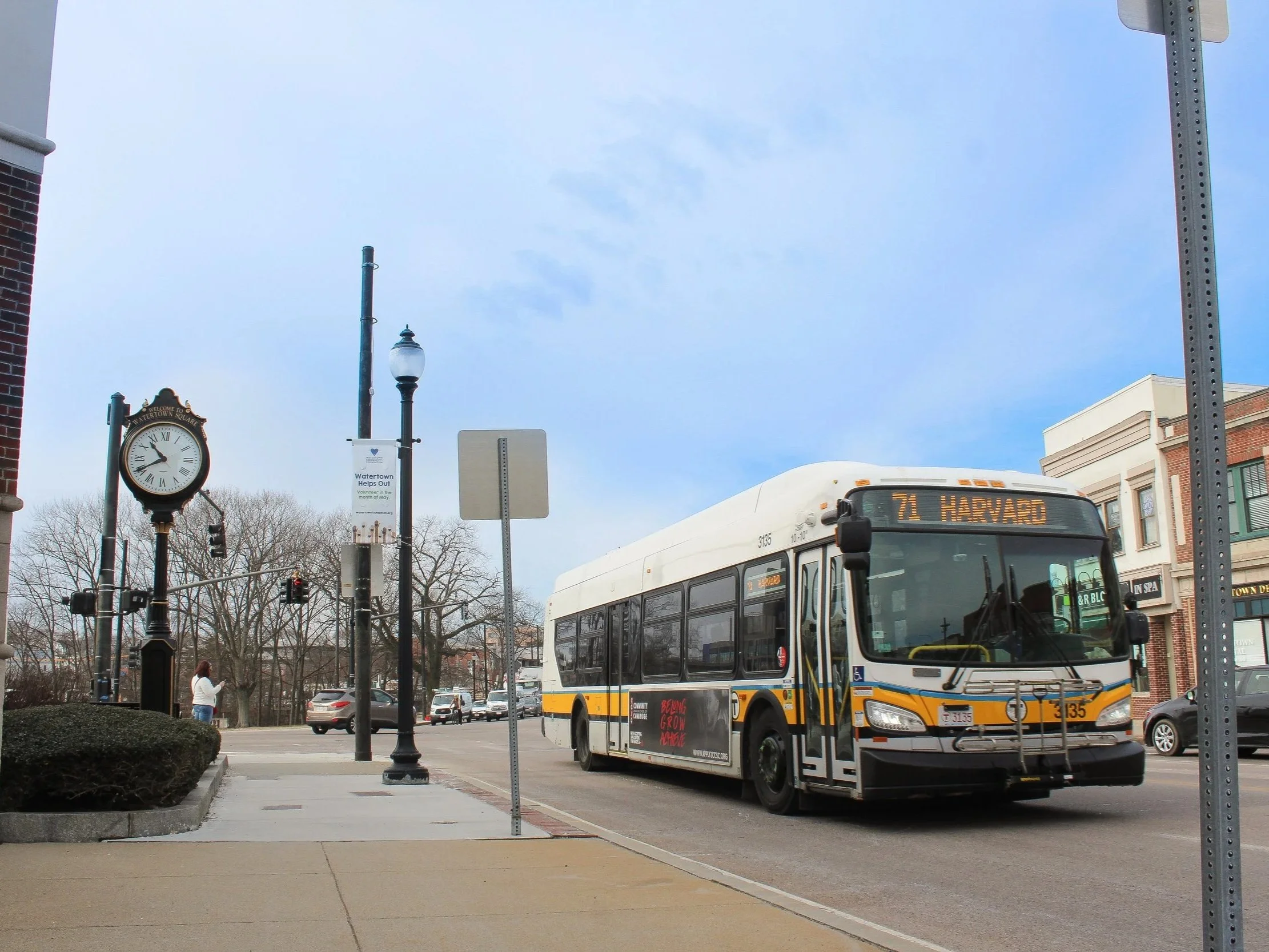 A public bus on a city street, displaying route 71 to Harvard, with a street clock, street lamps, trees, and buildings in the background.