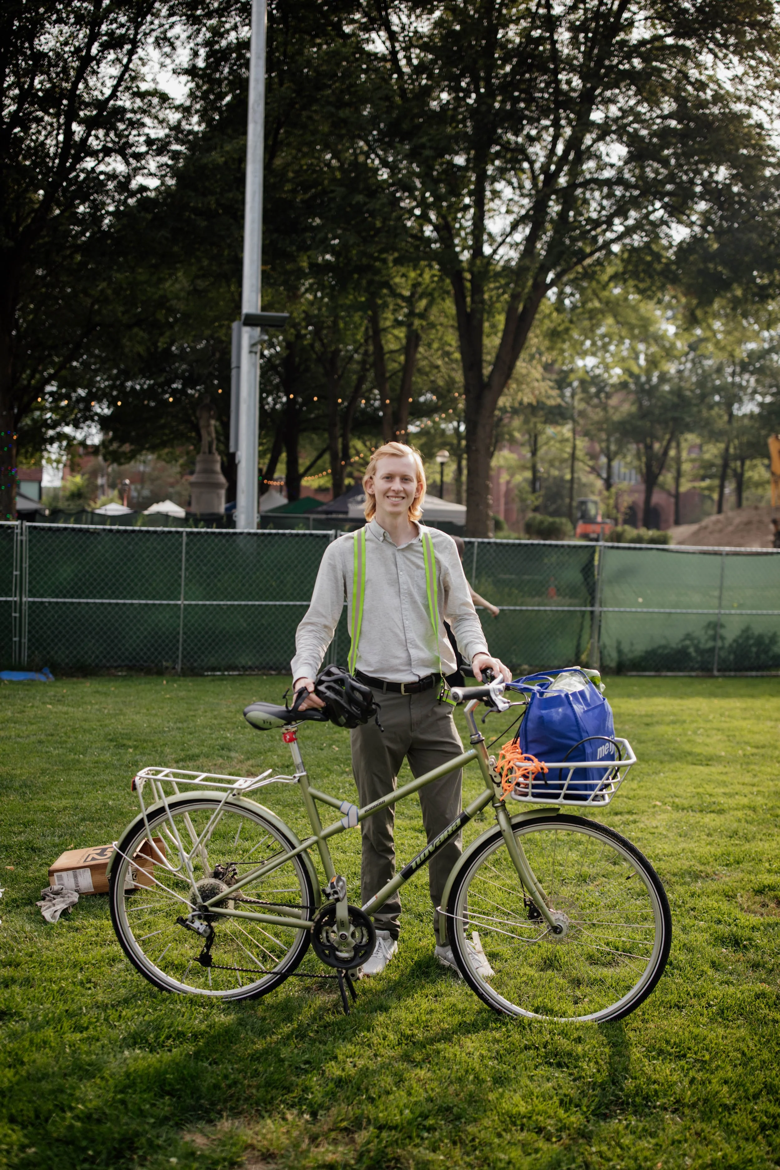 A young man in a light-colored shirt and gray pants stands outdoors with a bicycle, holding a helmet, in a park with trees and string lights in the background.