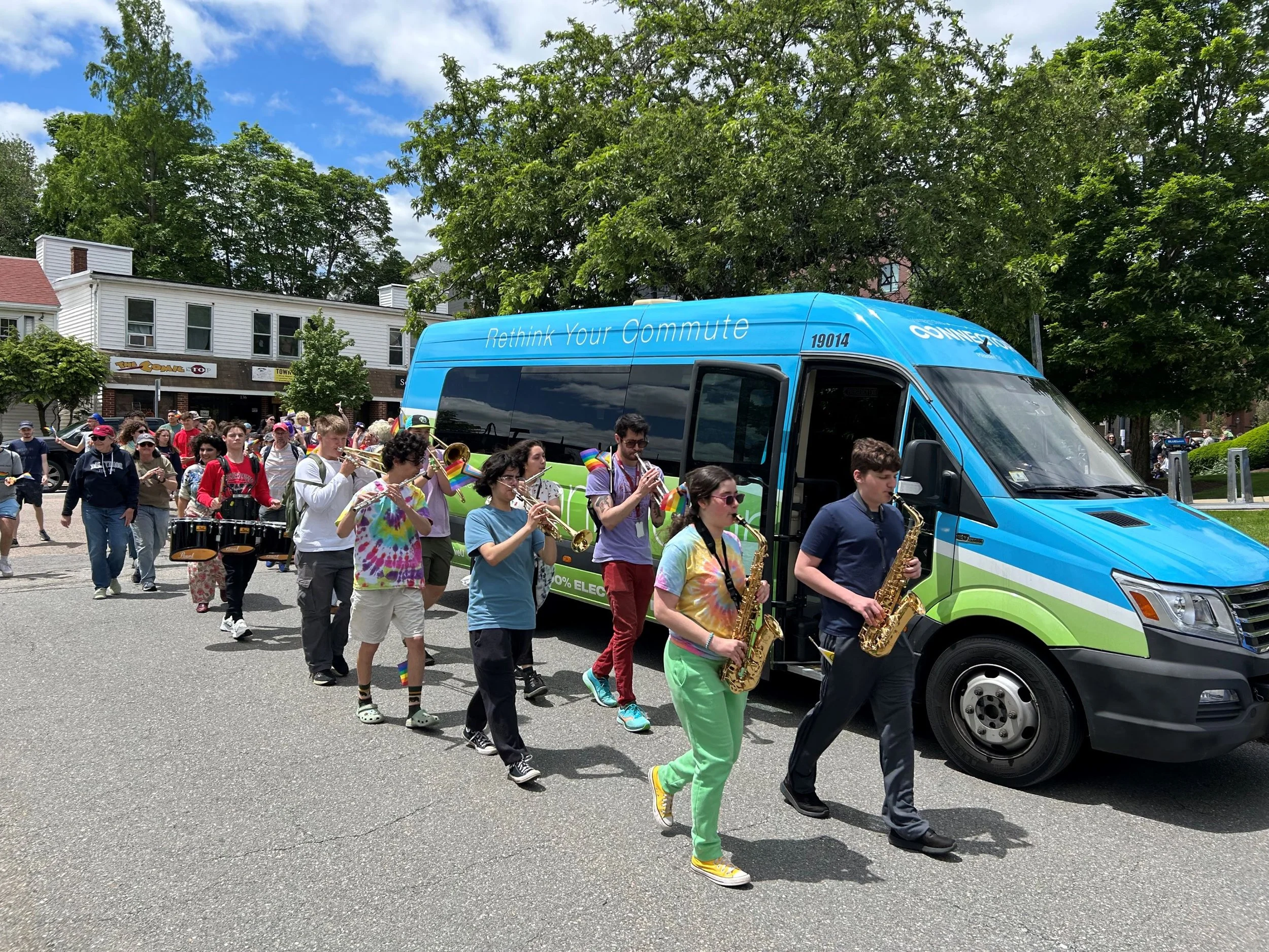 A parade featuring a marching band of young people playing instruments as they walk past a blue electric vehicle with 'Connect 4' and 'Rethink Your Commute' written on it, on a sunny day with people walking on the sidewalk and green trees in the background.