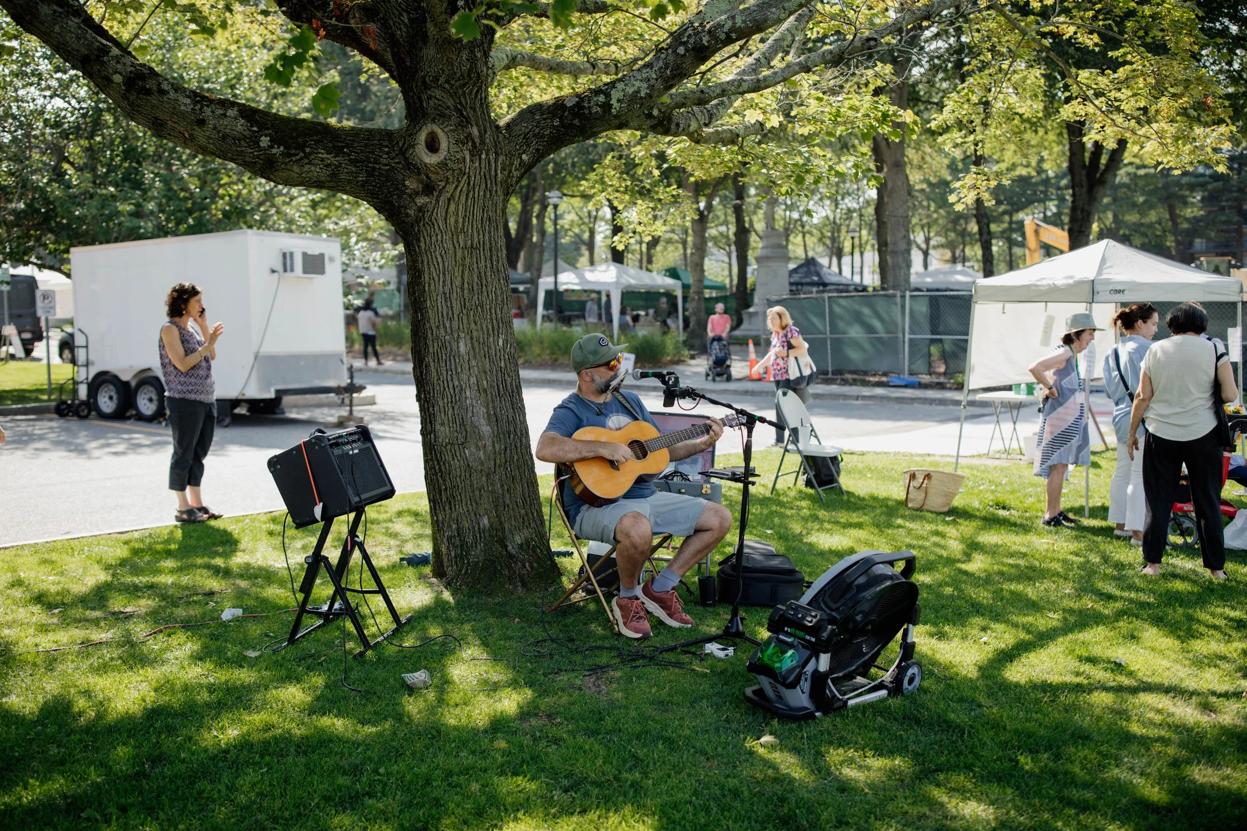A man playing a guitar and singing into a microphone under a large tree at an outdoor event, with people browsing booths and tents in the background.